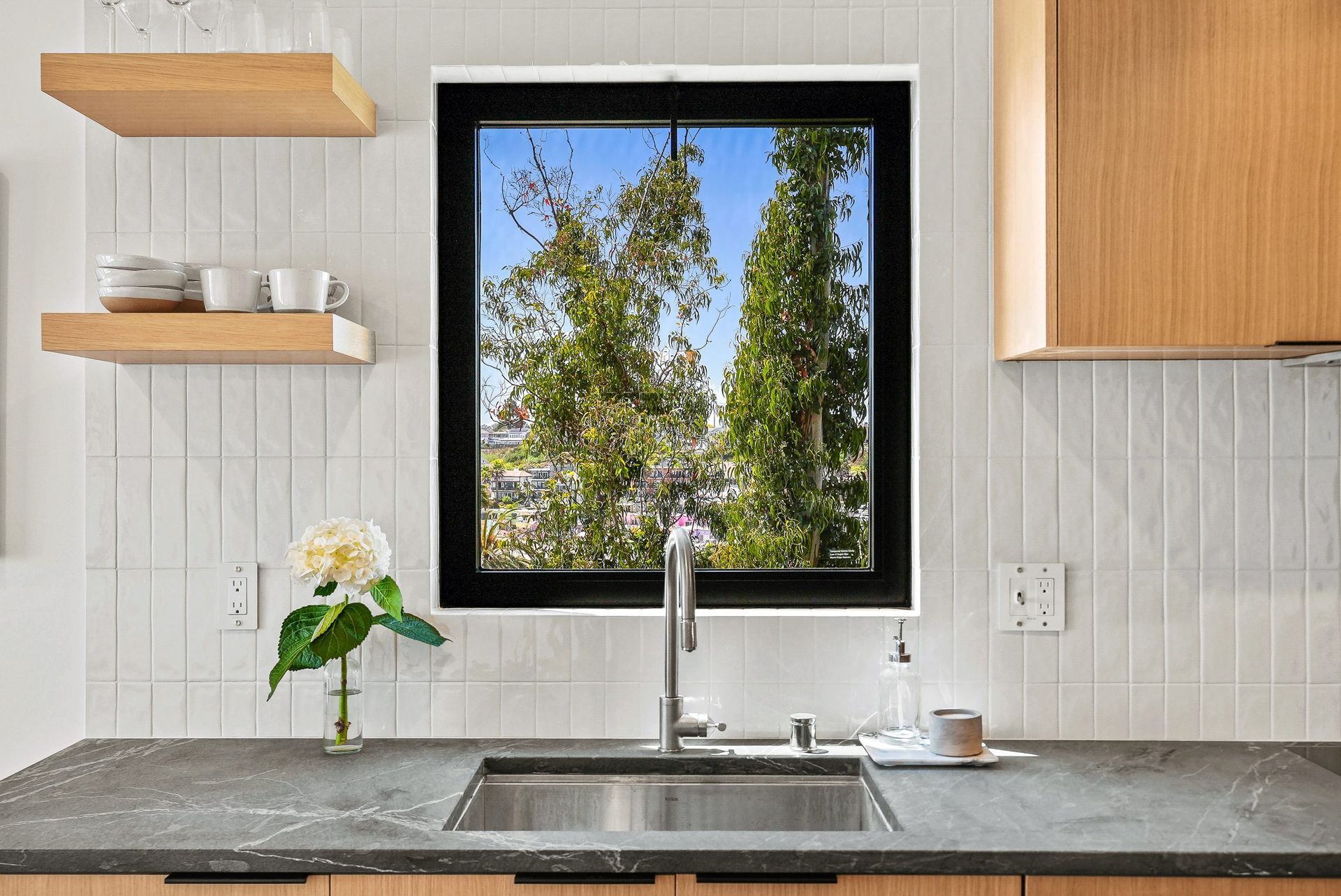 Modern kitchen sink beneath a window, with white tile, wood shelves, and a view of trees outside