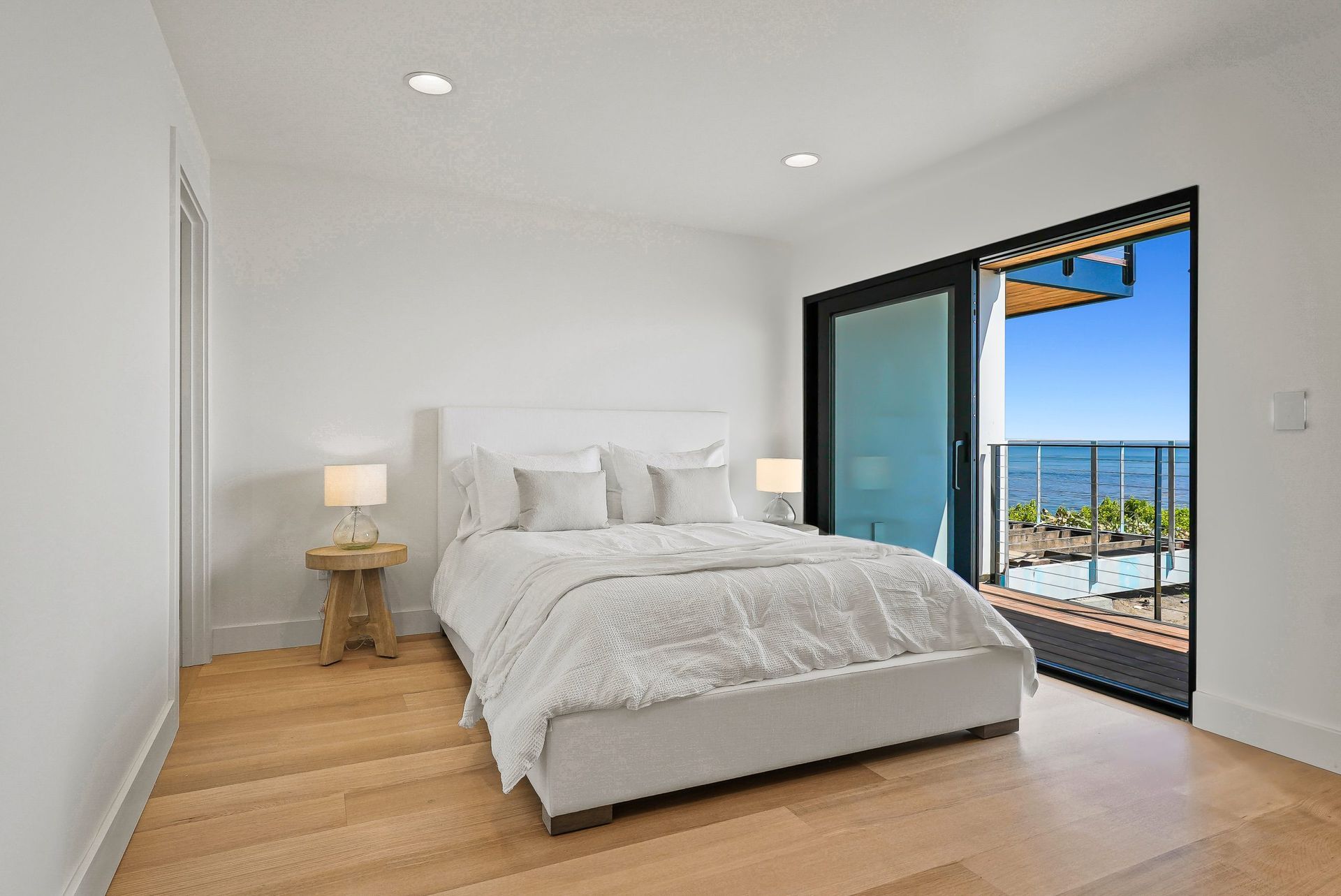 Bright minimalist bedroom with a bed, bedside lamp, and open balcony doors overlooking the ocean