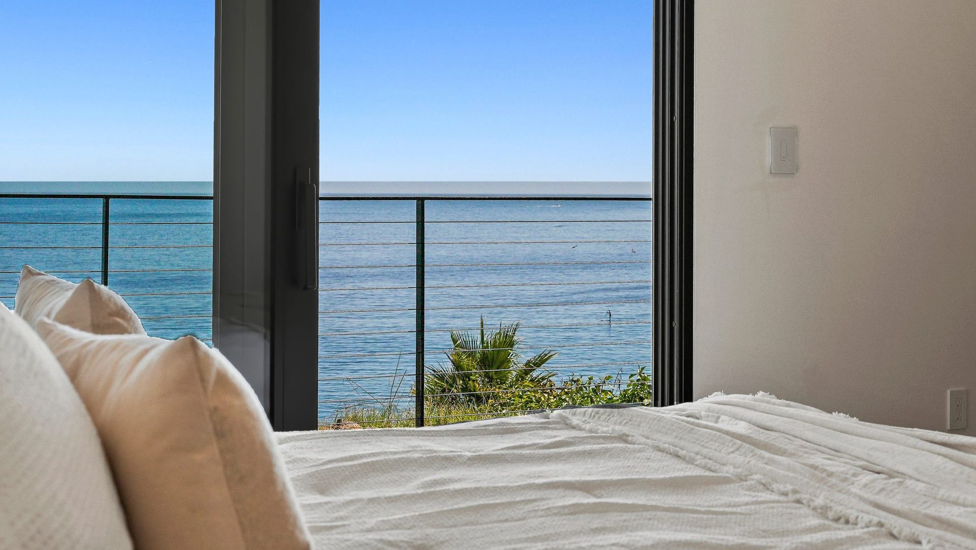 Bedroom with white bed linens overlooking a calm ocean through large windows at sunset