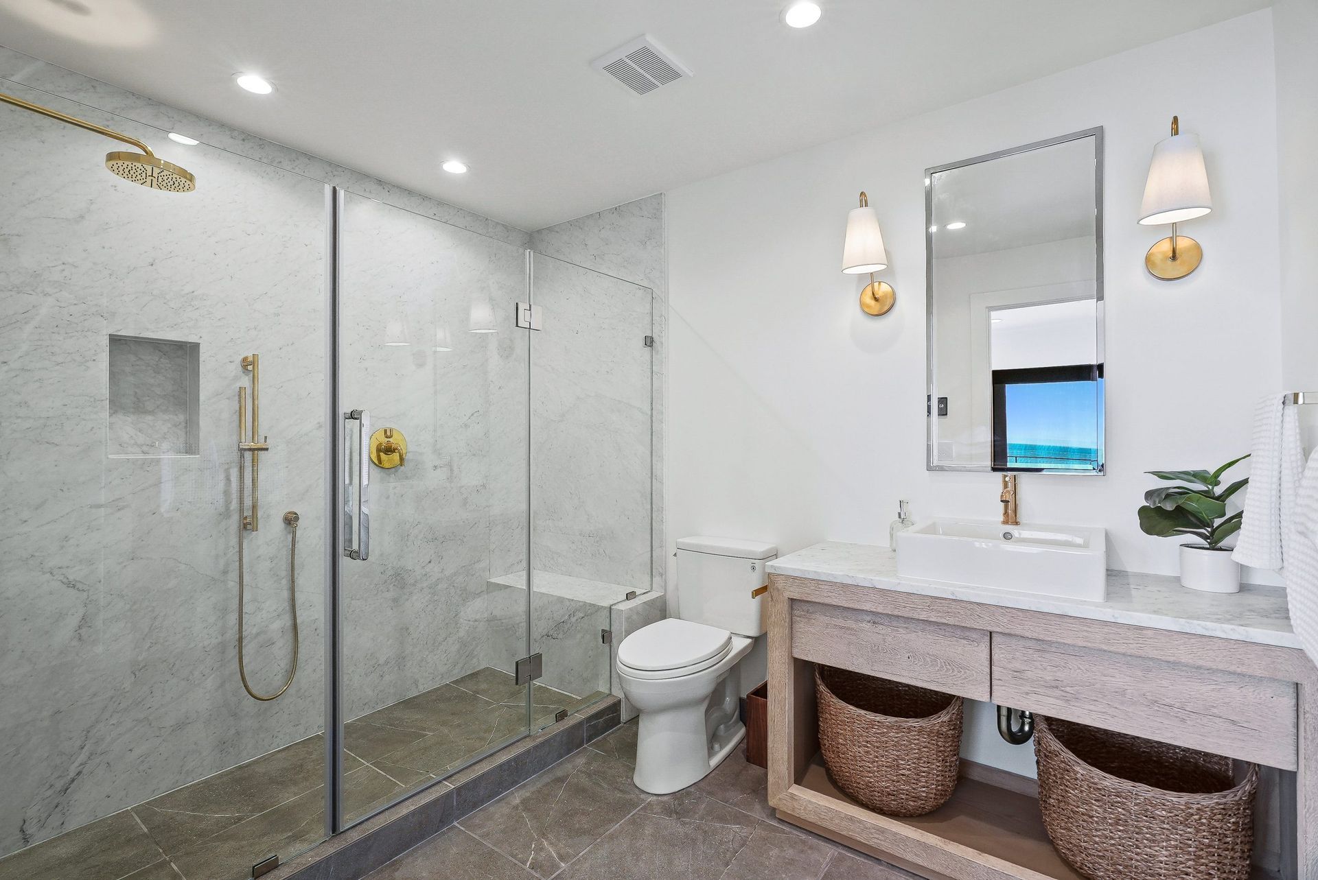 Bright modern bathroom with glass shower, toilet, vanity, mirror, and woven baskets underneath.