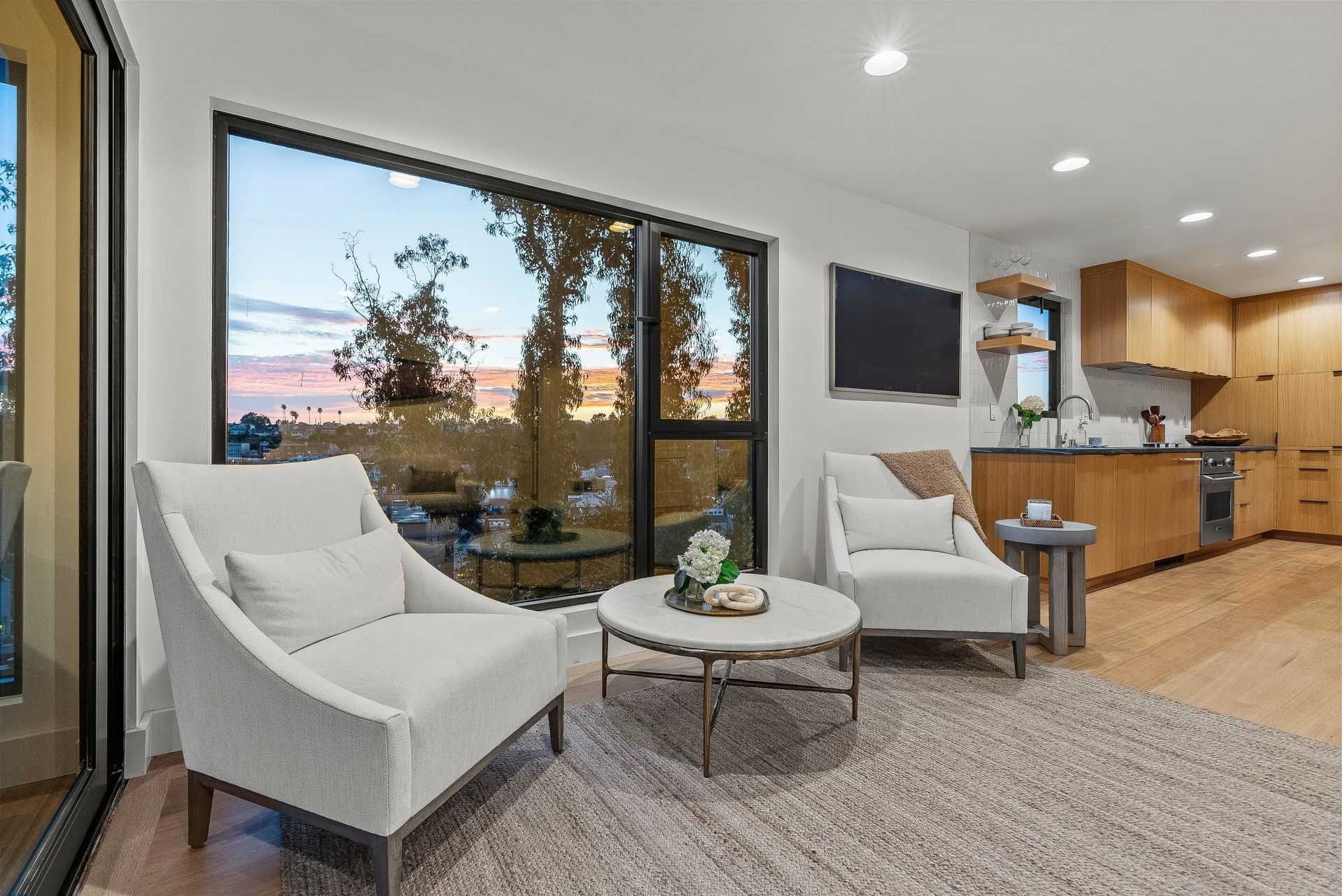 Modern living room with two white chairs, round coffee table, and kitchen area by large window
