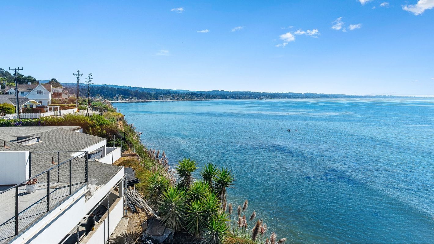 Sunny seaside coastline with blue water, rocky shore, and distant buildings under a clear sky