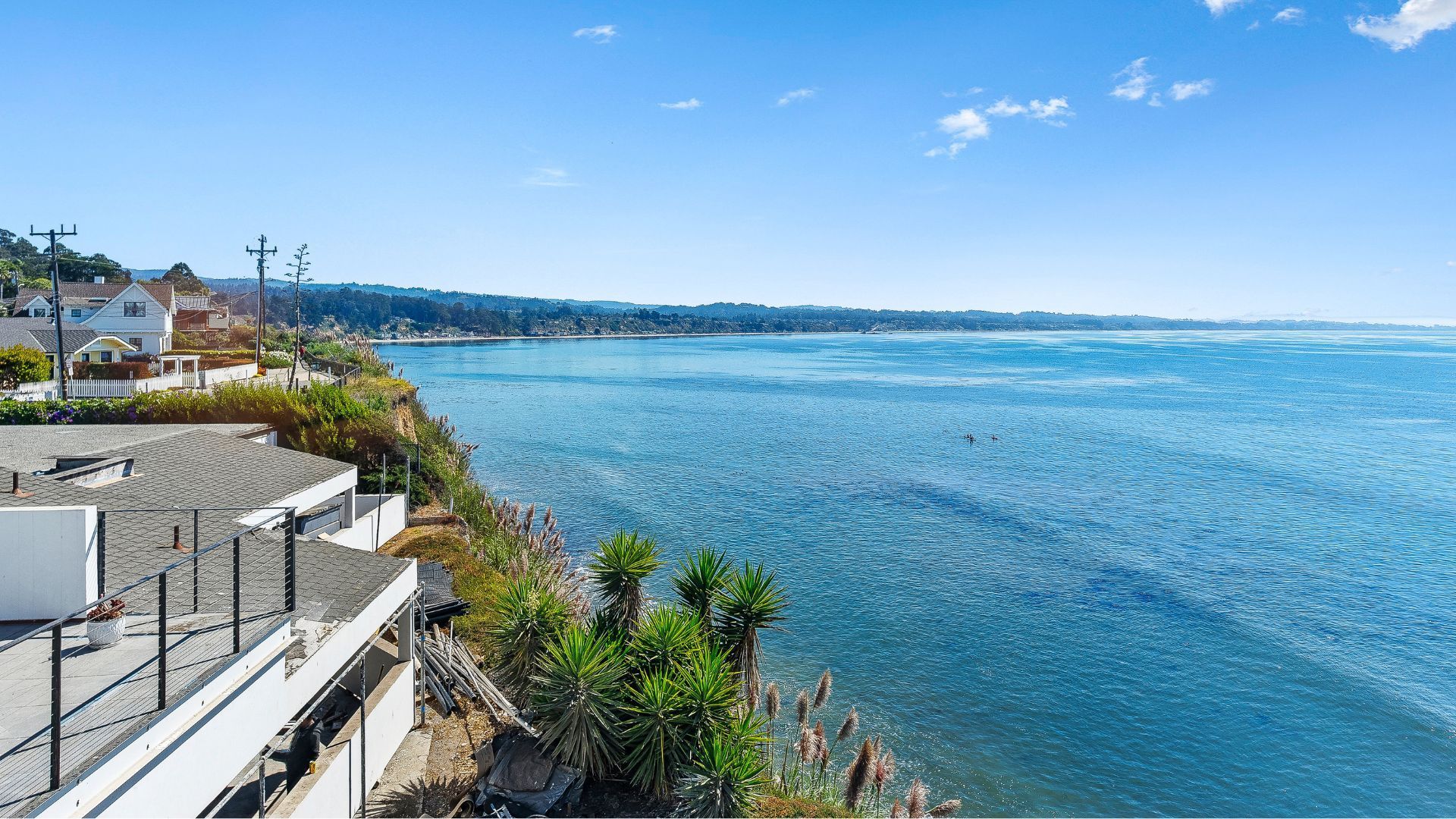 Coastal seaside view with blue water, rocky shore, and houses along the left side under a clear sky