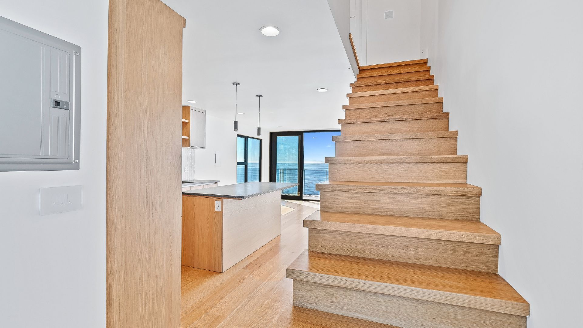 Bright modern home interior with wooden stairs, white kitchen, and large windows overlooking water
