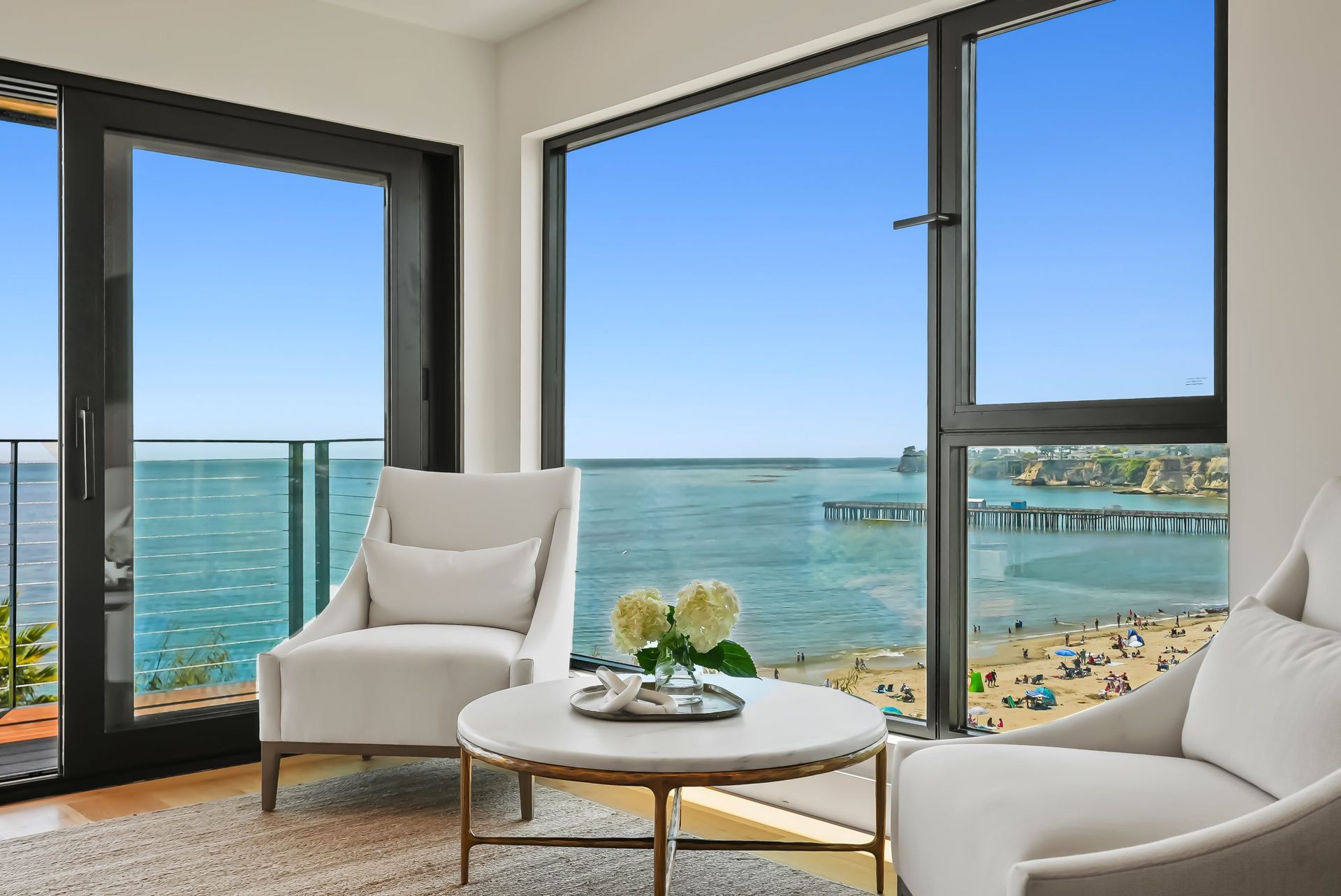Bright coastal living room with white chairs, round table, and large windows overlooking the ocean and beach.