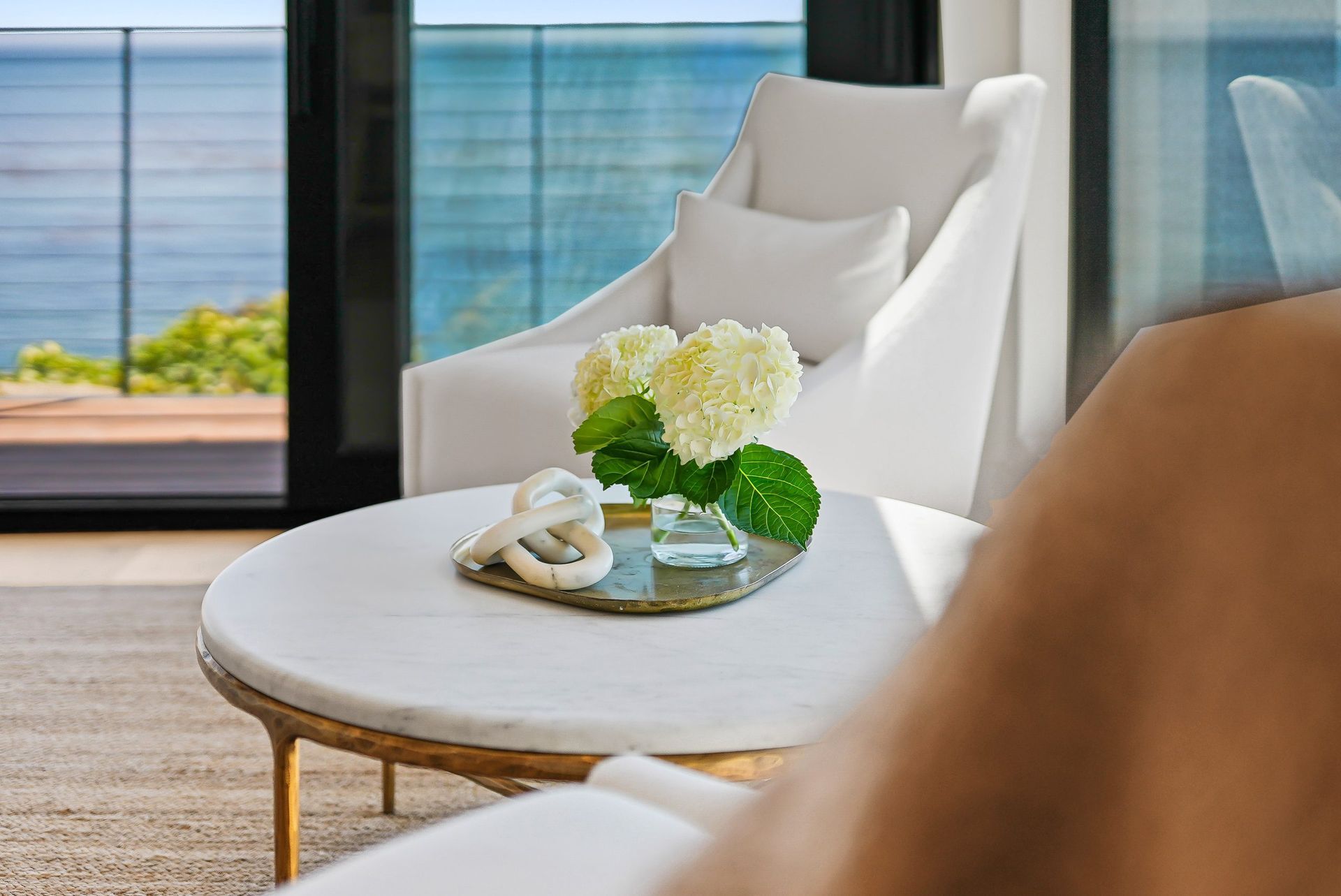 Bright living room with white chair, round table, flowers, and ocean view through large windows