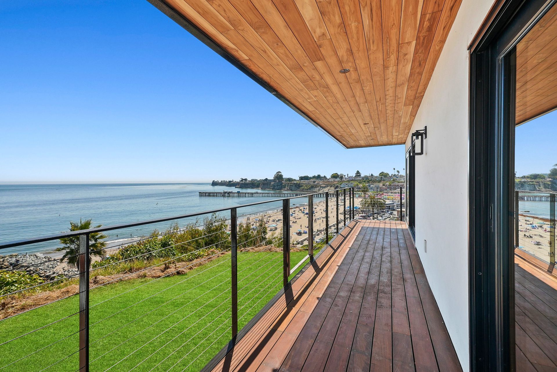 Oceanfront deck with black railing, wooden ceiling, and view of water and shoreline under blue sky