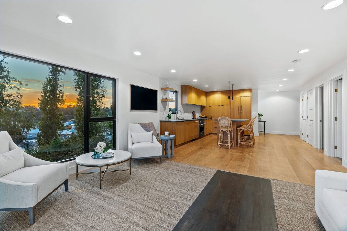Bright open-concept living room with large windows, gray seating, and a kitchen with wood cabinets and island.