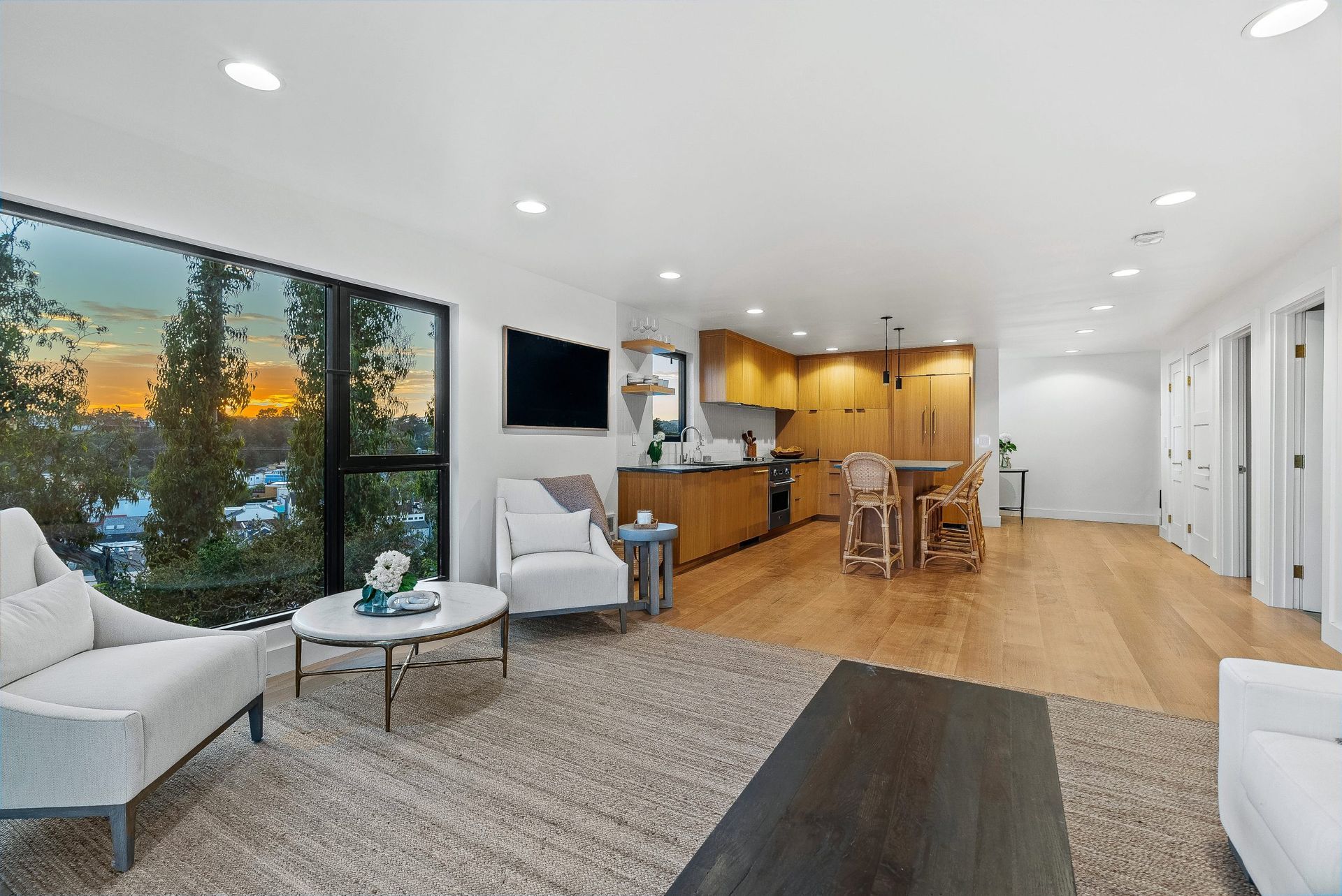 Bright open-plan living room with large windows, gray chairs, rug, and kitchen with wood cabinets at the back.