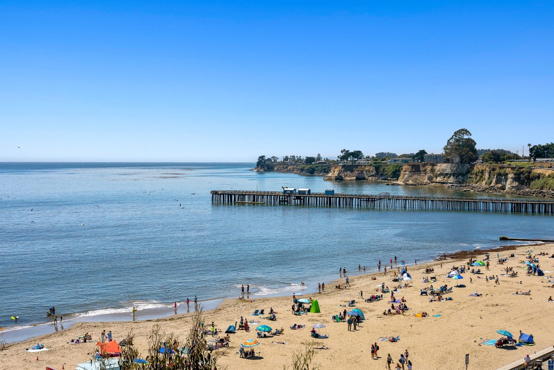 Sunny beach with people, calm blue water, and a pier stretching into the sea.