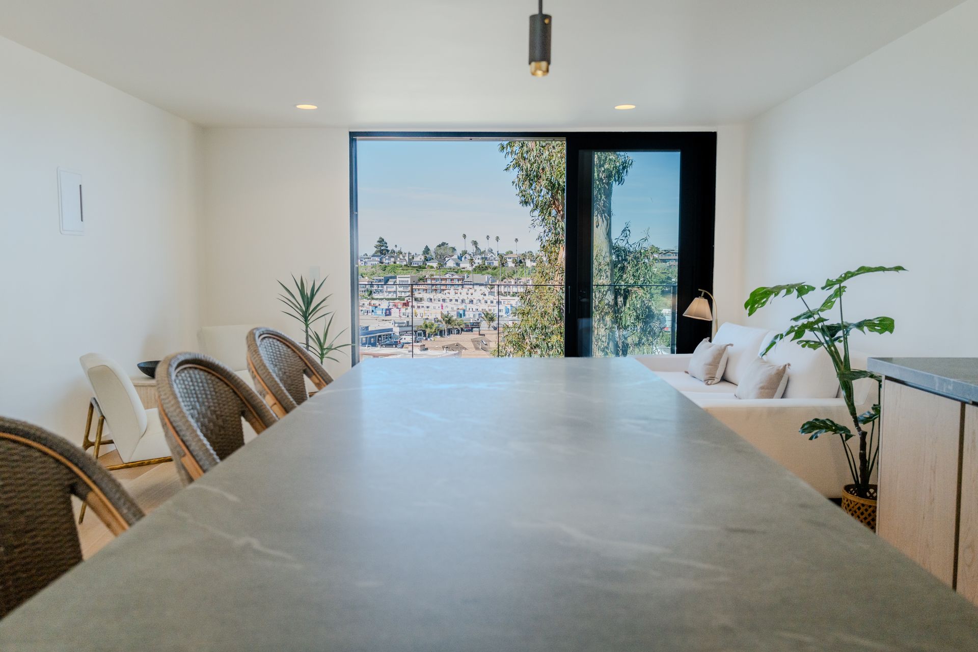 Modern dining room with a long stone table, woven chairs, and a large window overlooking a city view