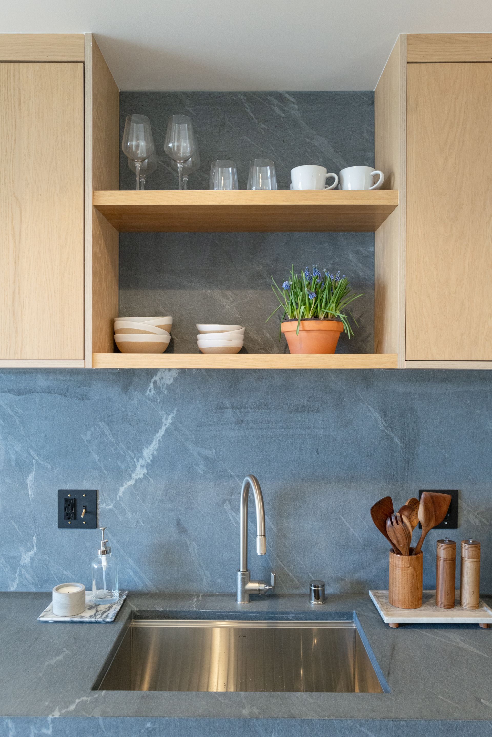 Modern kitchen sink with stainless basin, blue backsplash, open shelves, dishes, glasses, plant, and utensils