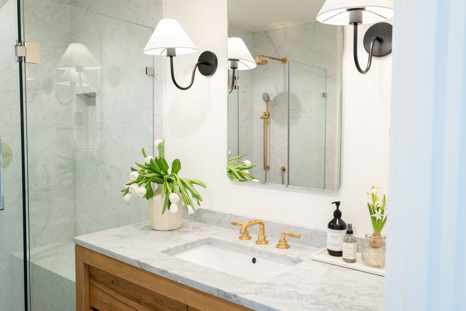 Bright bathroom vanity with marble countertop, gold faucet, mirror, wall sconces, and potted plants