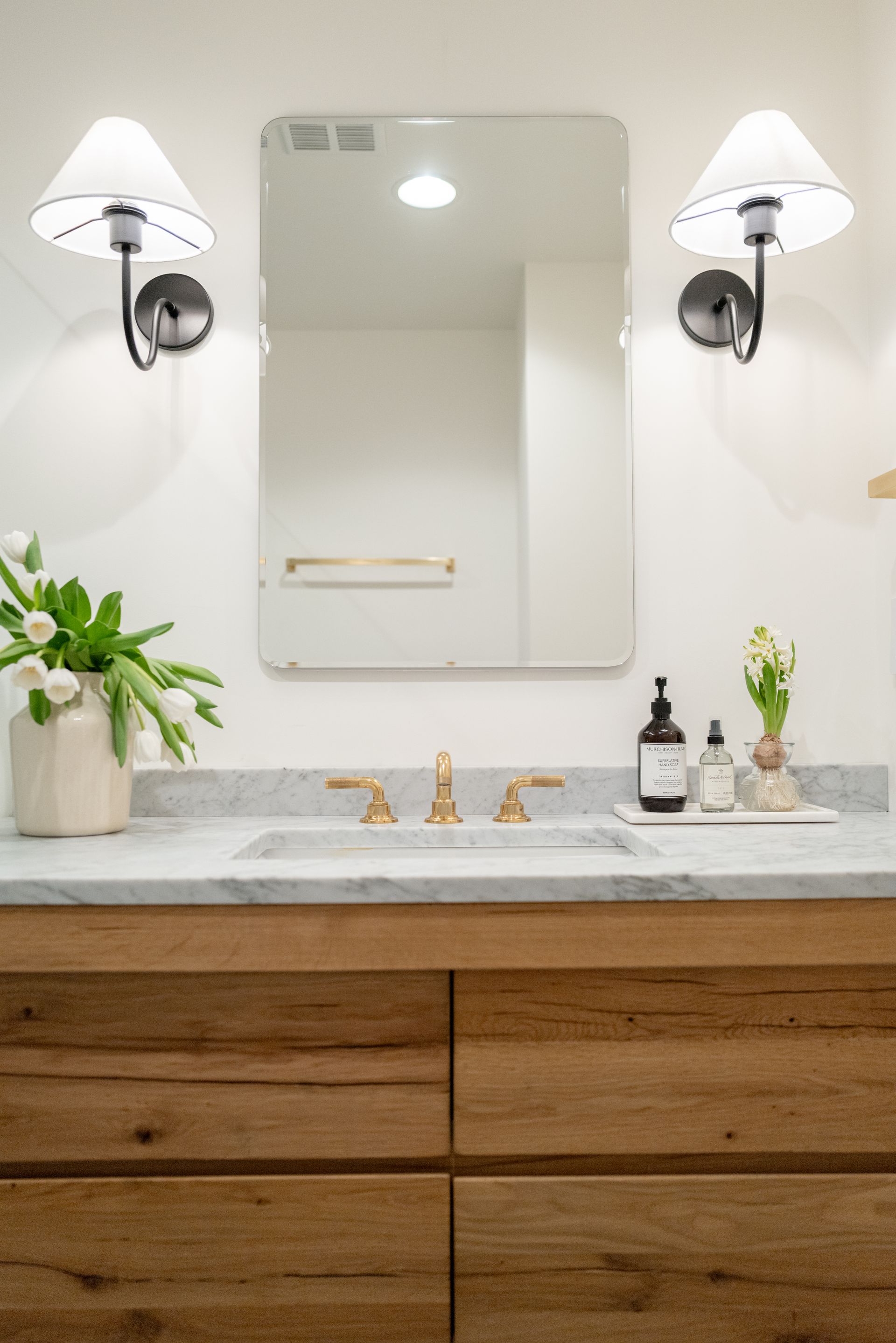 Modern bathroom vanity with mirror, wall sconces, white countertop, wood cabinets, and potted plants