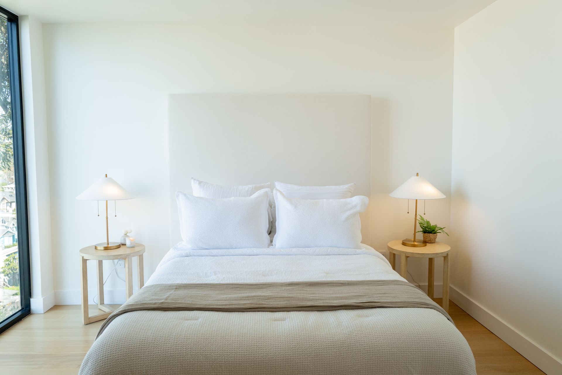 Bright minimalist bedroom with a white bed, bedside lamps, and large windows on the left.