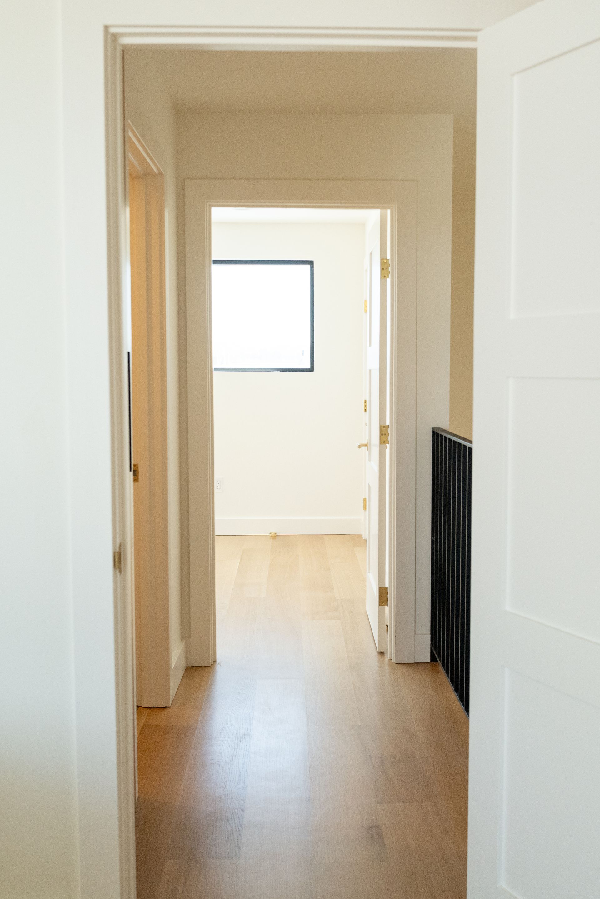 Narrow hallway with light wood floor, white walls, and a bright room visible at the end