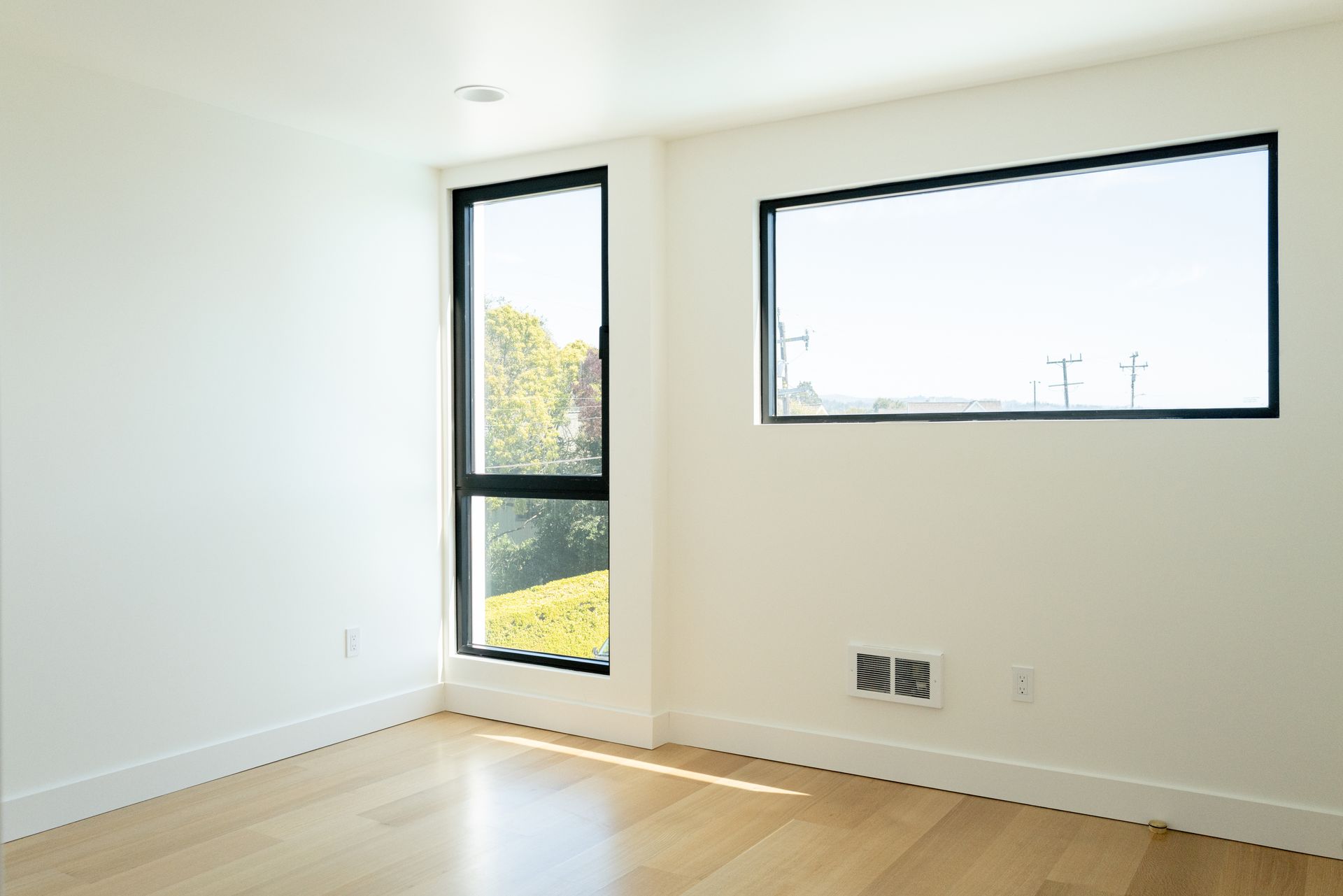 Bright empty room with light wood floor, white walls, and two black-framed windows.