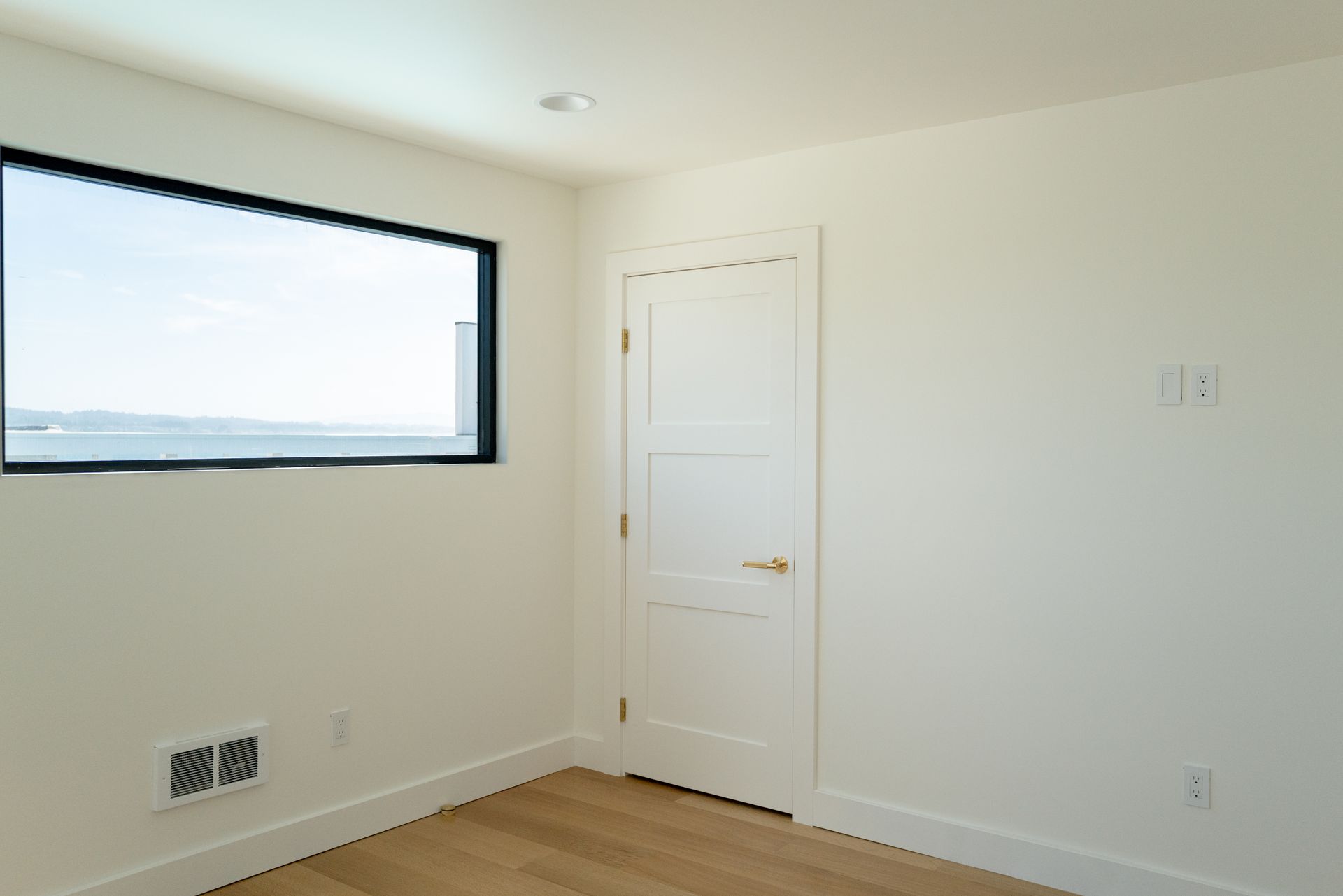 Empty white room with a window, closed door, and wood floor.