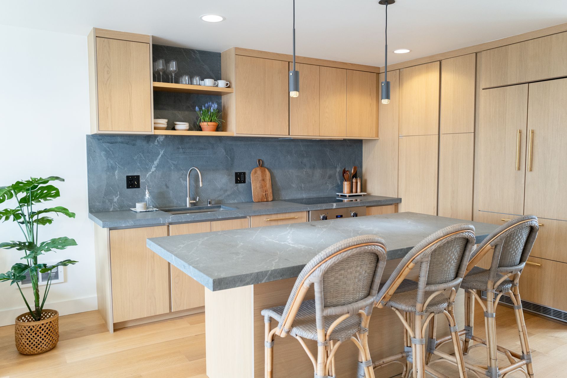Modern kitchen with light wood cabinets, gray island, woven bar stools, and a potted plant