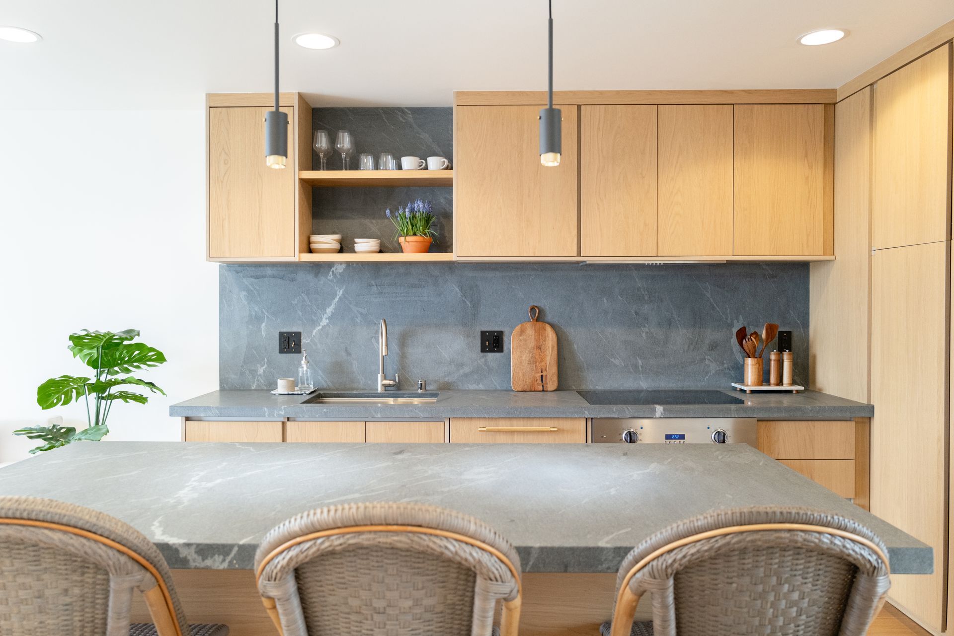 Modern kitchen with light wood cabinets, gray backsplash, and a counter with woven stools in front.