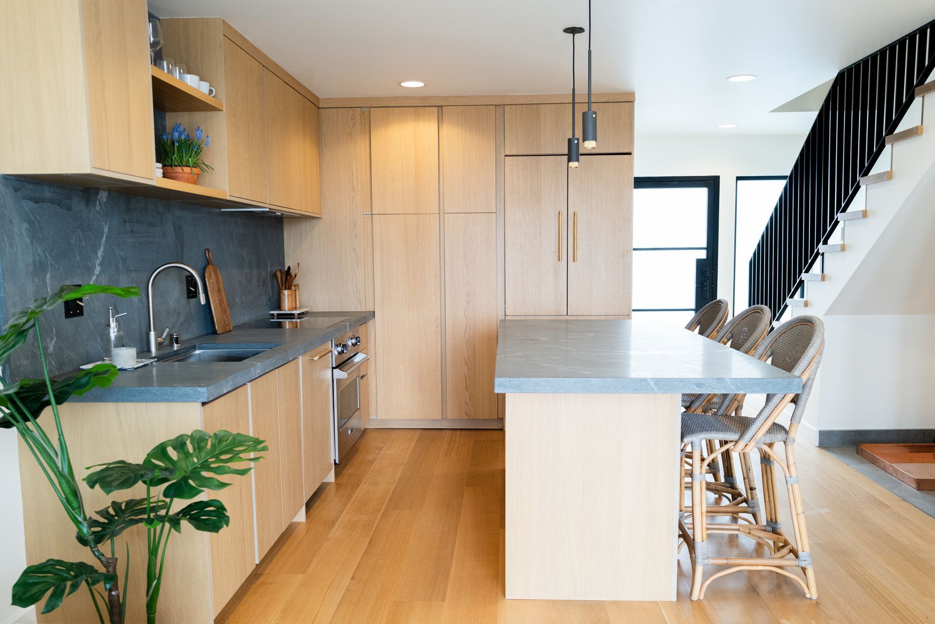 Bright modern kitchen with wood cabinets, gray countertops, island, and black staircase beside dining chairs