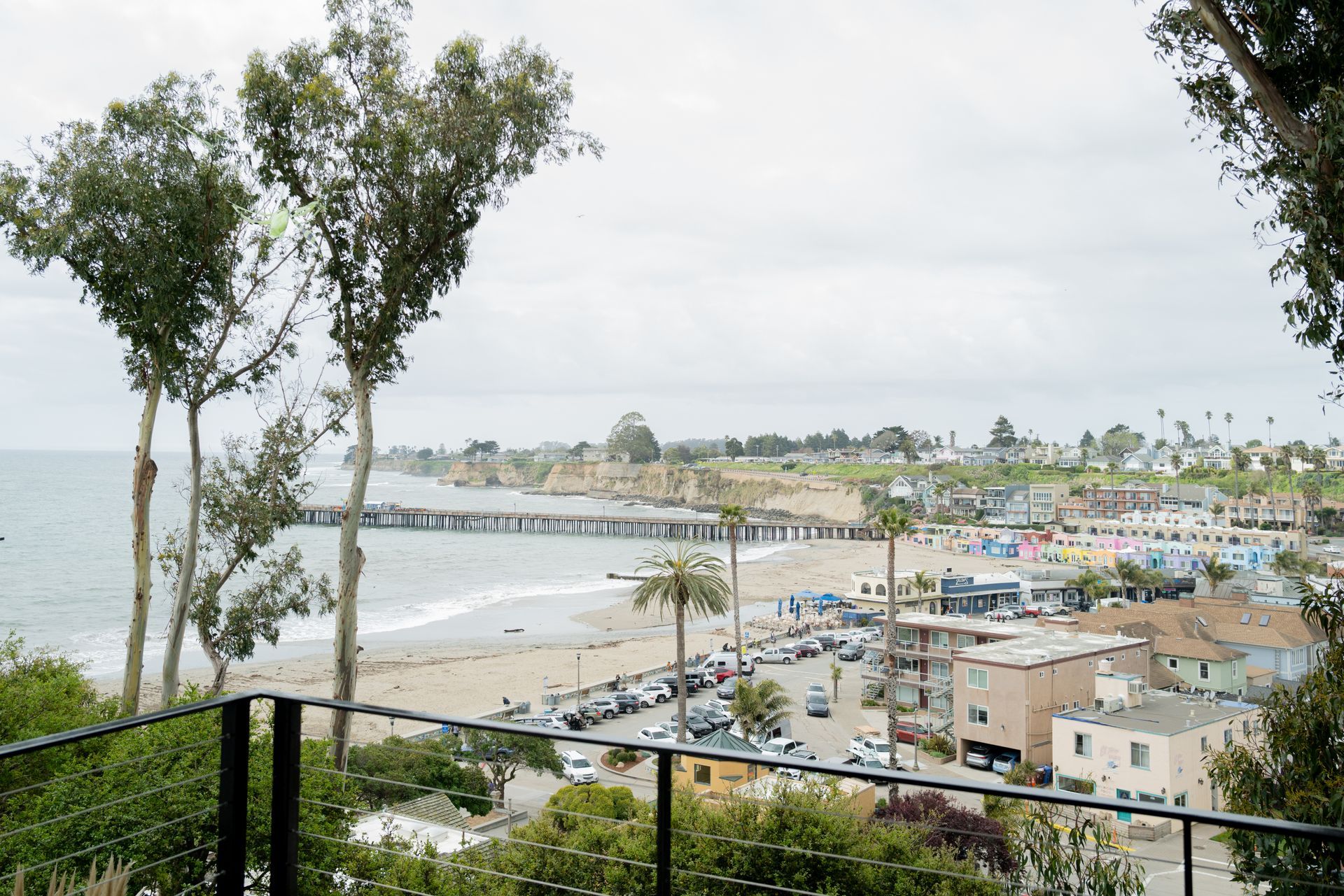 Coastal bay with beach, pier, and town buildings under a cloudy sky, viewed from a hilltop balcony