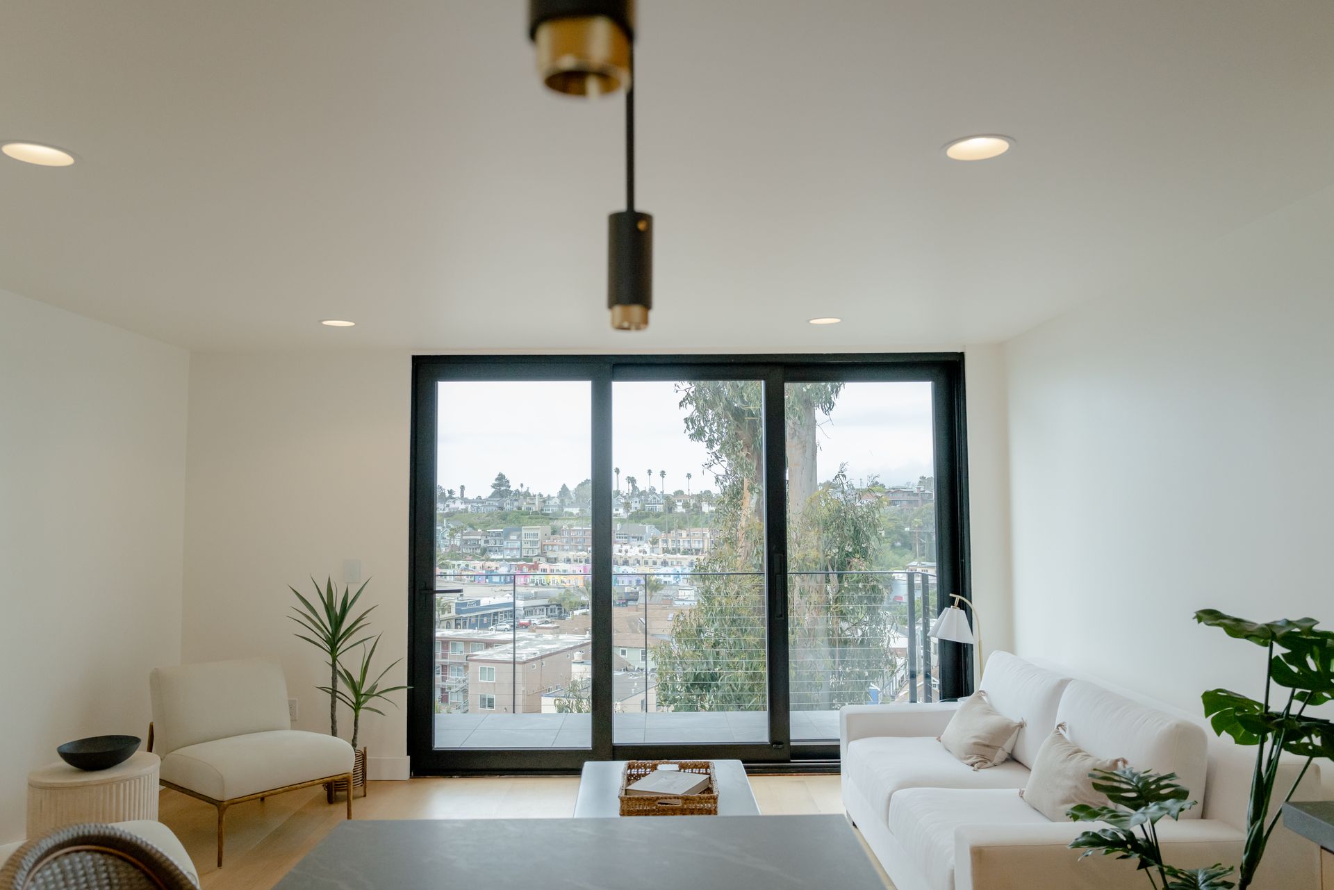 Bright modern living room with white sofa, plants, and large black-framed windows overlooking a city view