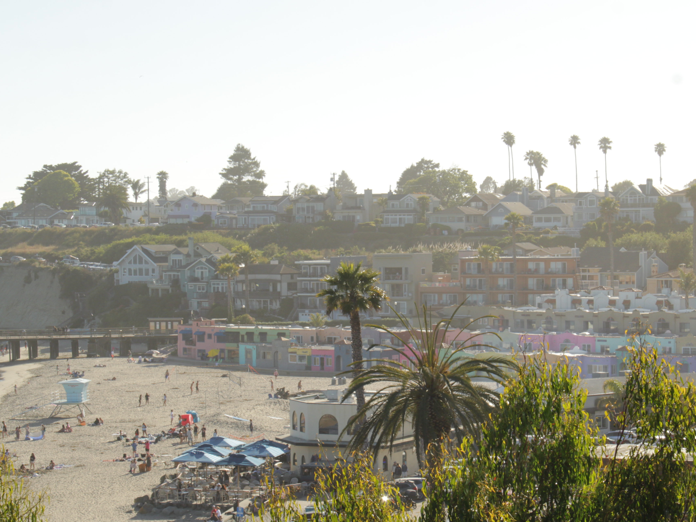 Crowded sunny beach with umbrellas, palm trees, and hillside houses in the background