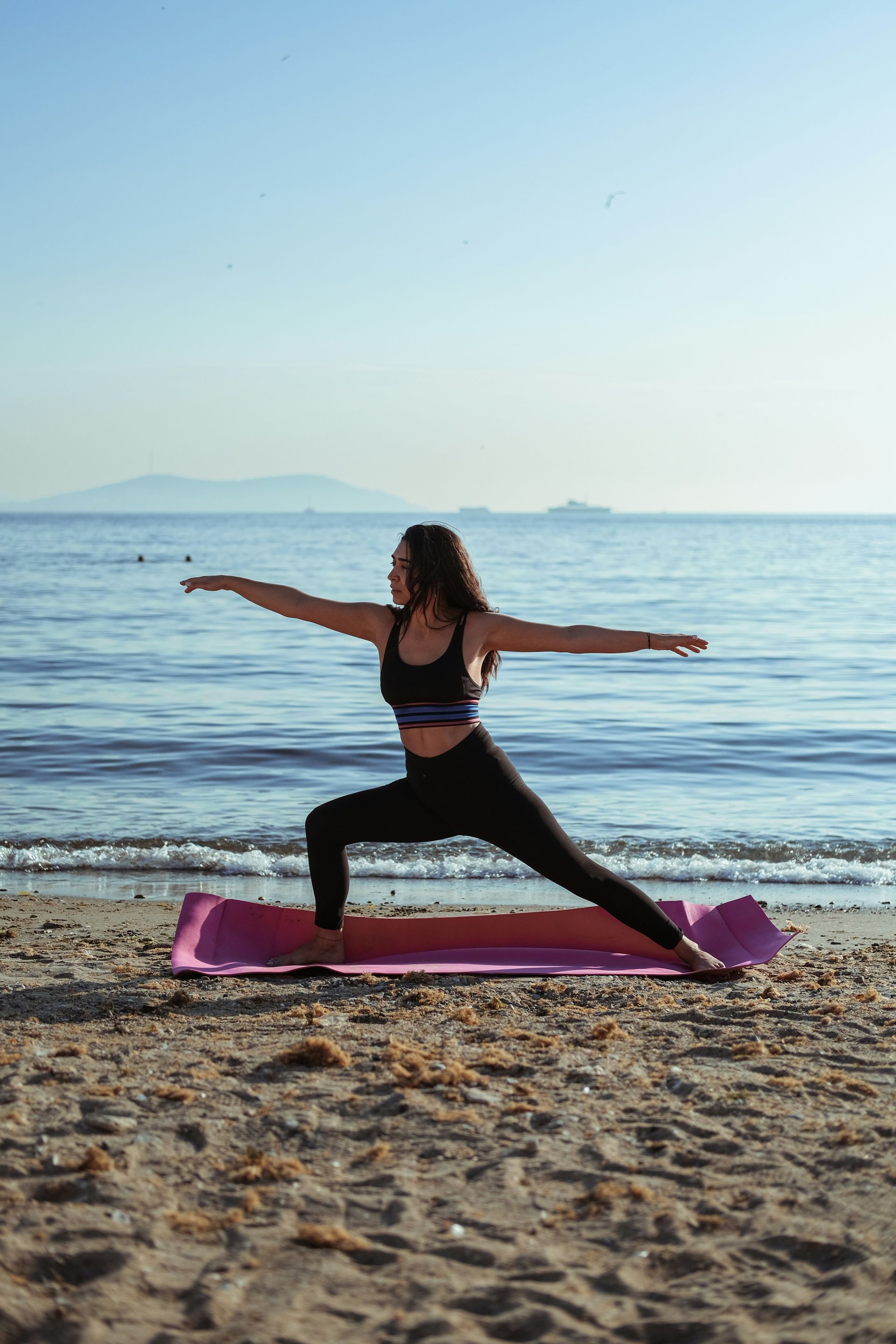 Person doing a yoga warrior pose on a pink mat at the beach at sunset
