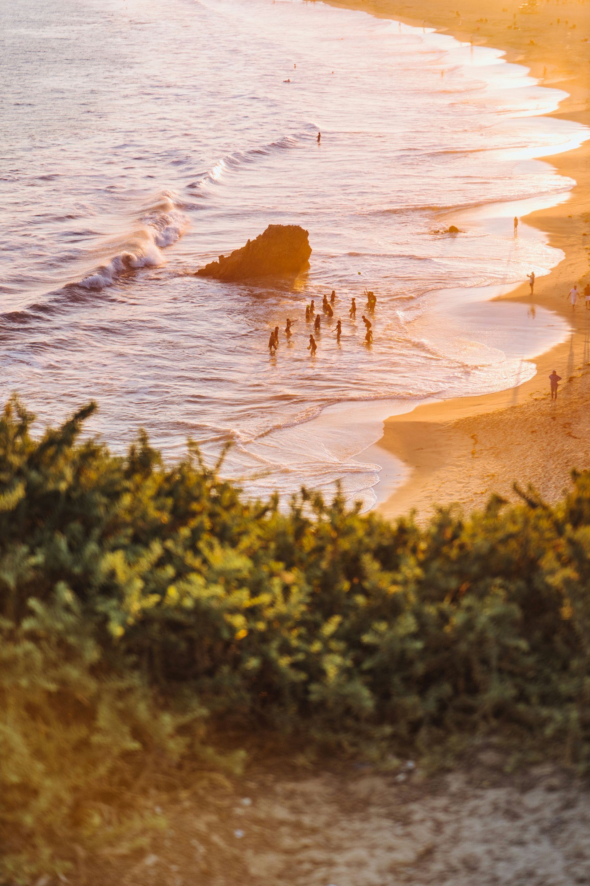 Sunset beach with gentle waves, a rock in the surf, and a few people standing near the shoreline.