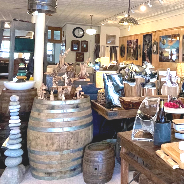 Interior of a retail shop displays wooden furniture, art, and home decor; wine barrels and stone stacks are prominent.