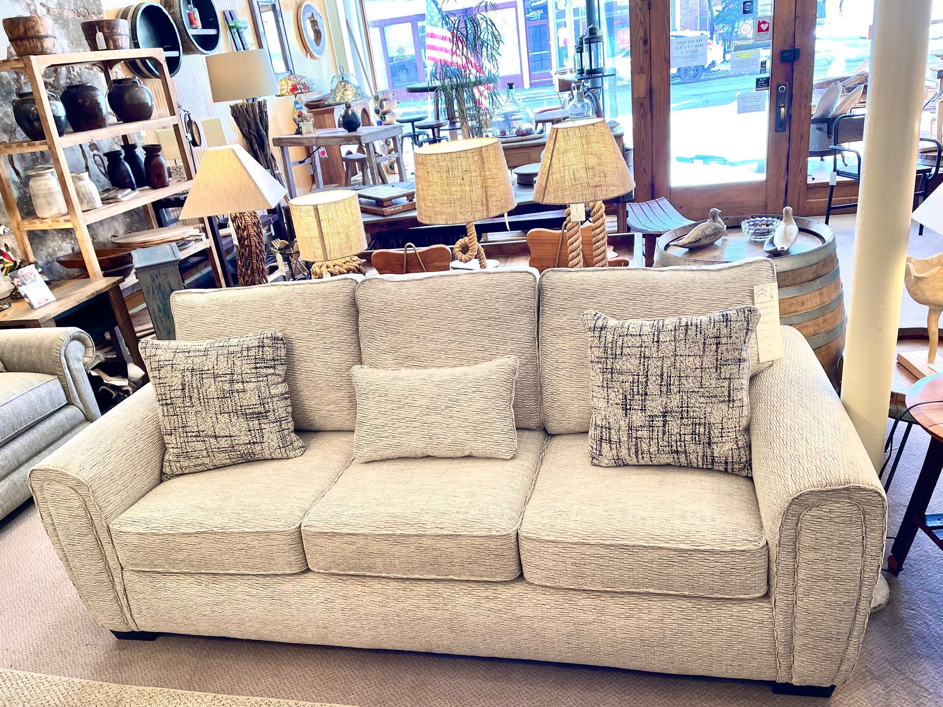 Cream-colored couch with patterned pillows in a furniture store. Lamps, shelves, and a barrel are in the background.