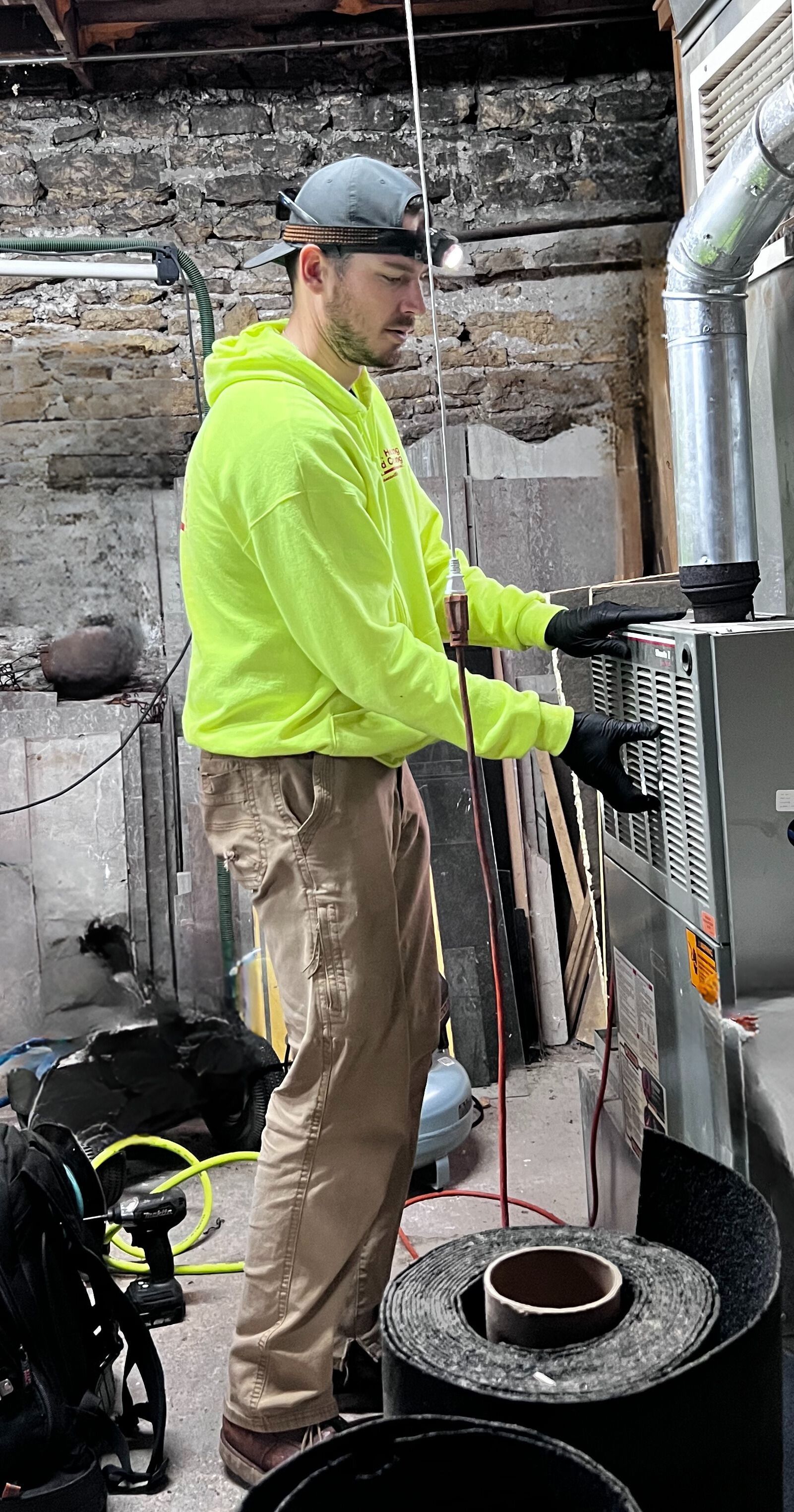 Worker in neon yellow hoodie using a tool in a basement utility room next to HVAC equipment.