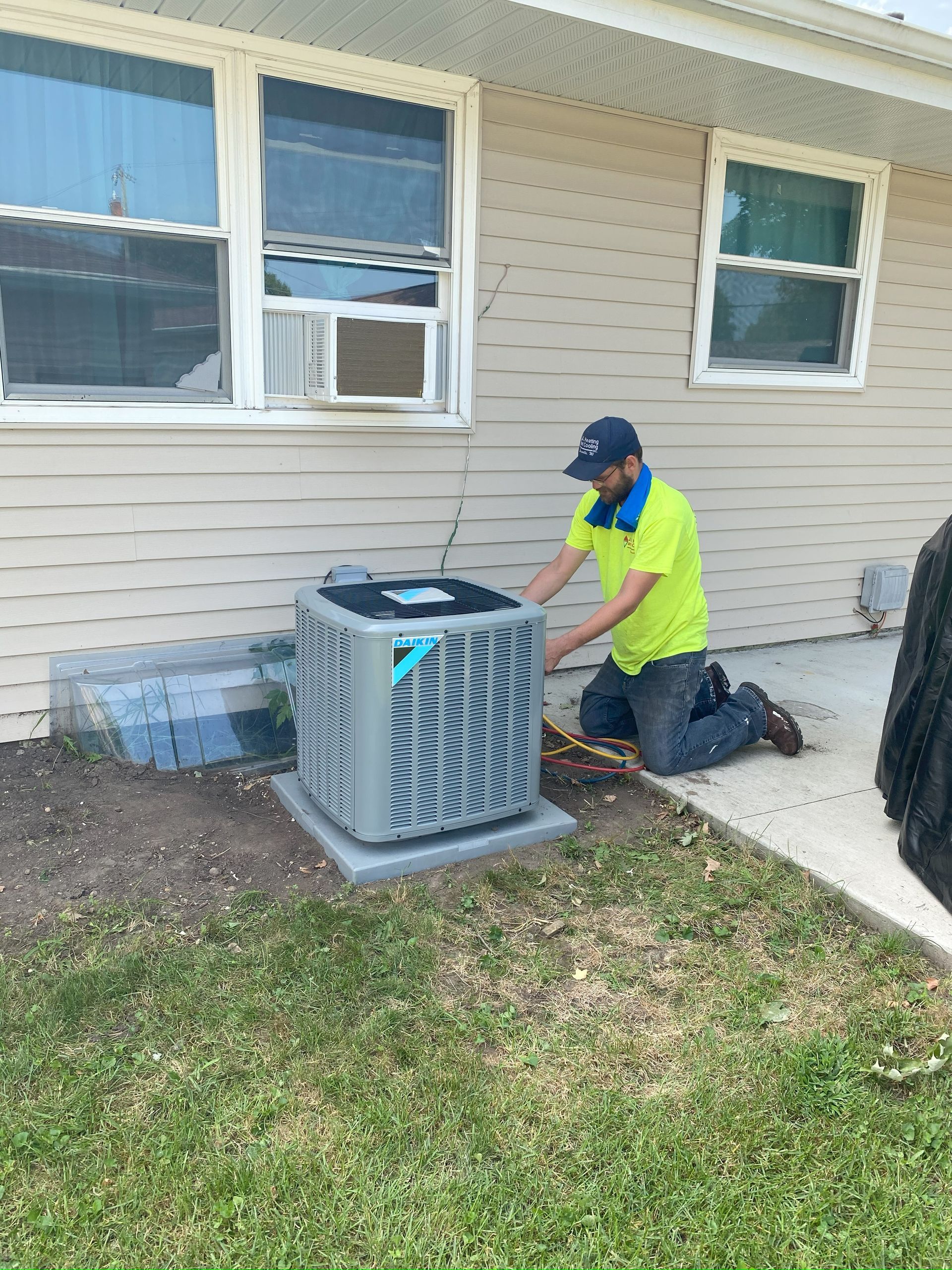 Worker kneeling beside a house, adjusting an outdoor air unit near two windows.