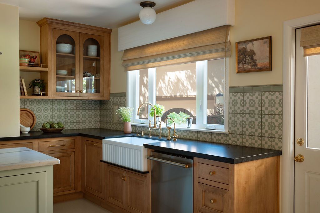 A cozy kitchen featuring light wood cabinets, a farmhouse sink, patterned backsplash tiles, and a window with a blind.