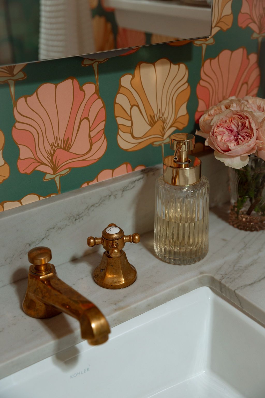 Close-up of a white sink with gold faucet, floral wallpaper, and pink flowers.