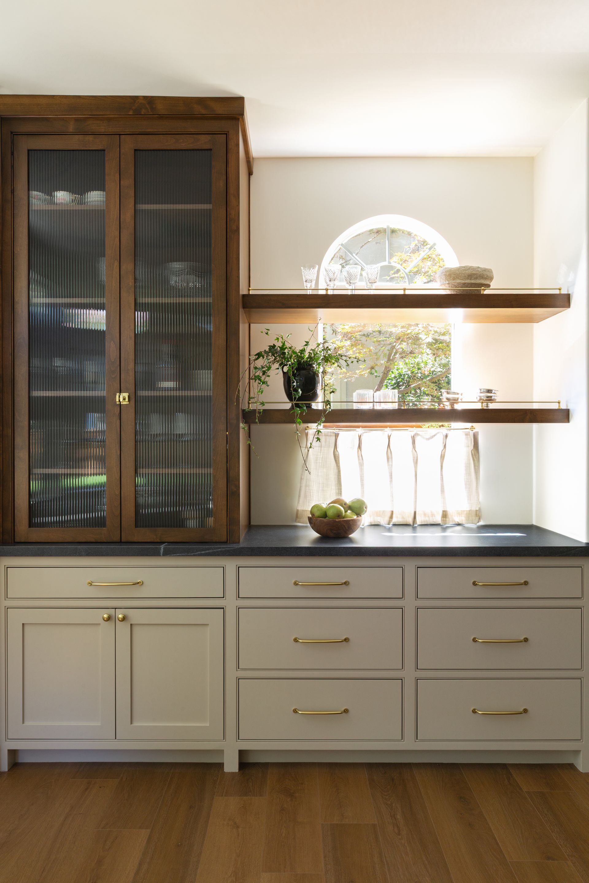 A wooden cabinet with reeded glass doors sits next to shelves and light gray cabinets with gold hardware in a kitchen.
