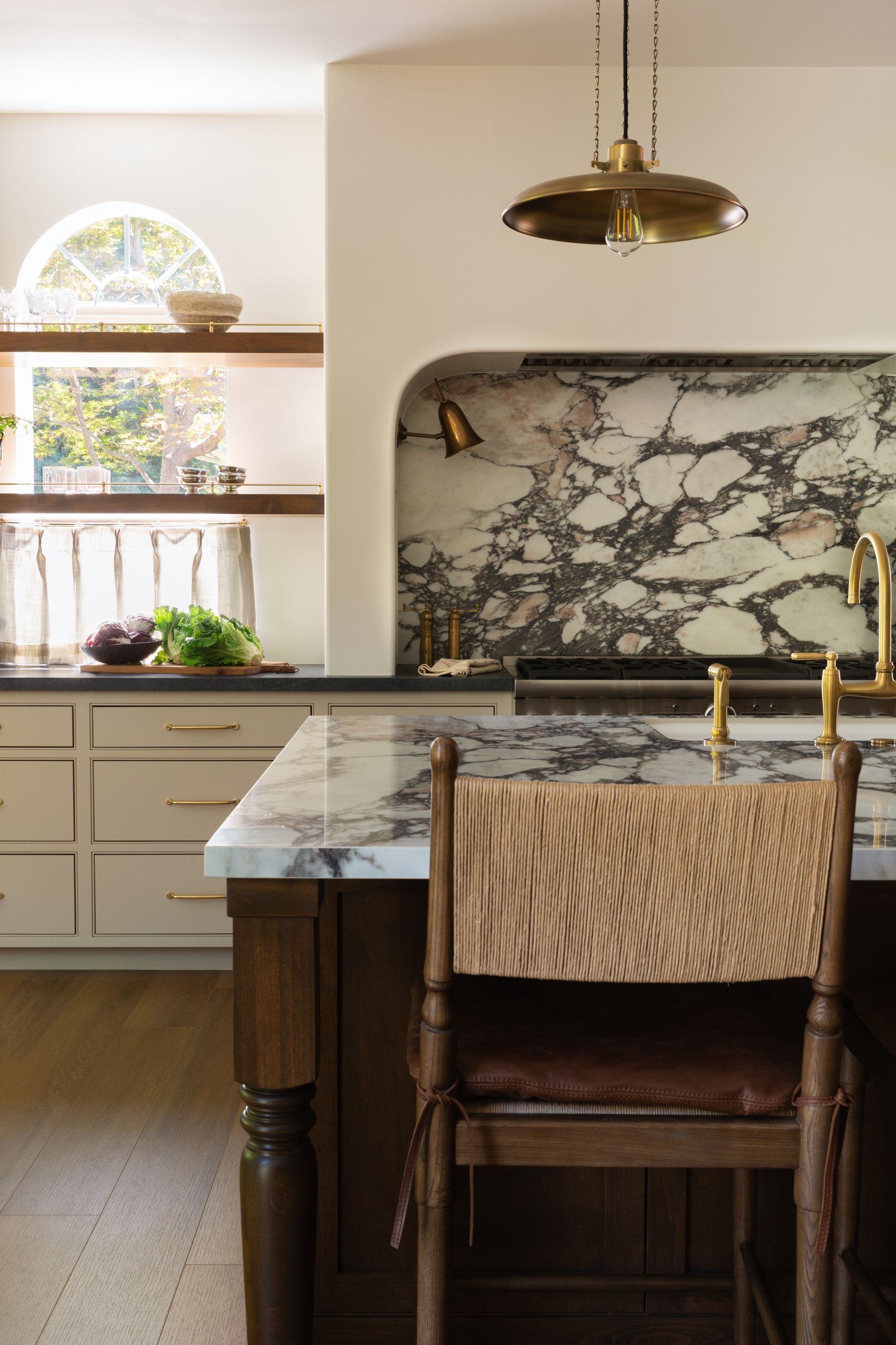 A kitchen featuring a marble-topped island with a wooden stool, a marble backsplash in an arched alcove, and open shelves.