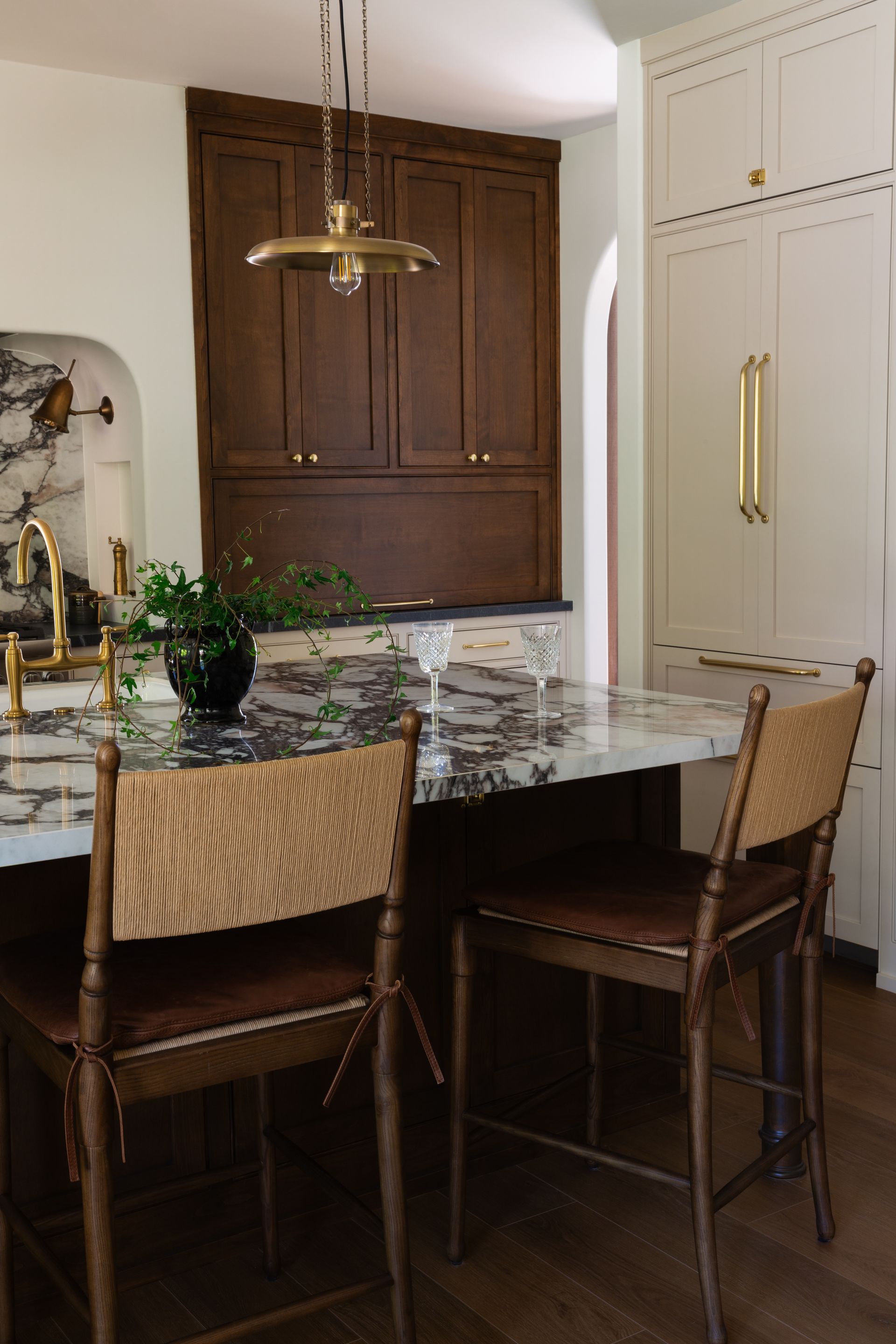 Luxury kitchen remodel with cane-back stools, a marble island, dark wood cabinetry, and a brass pendant.