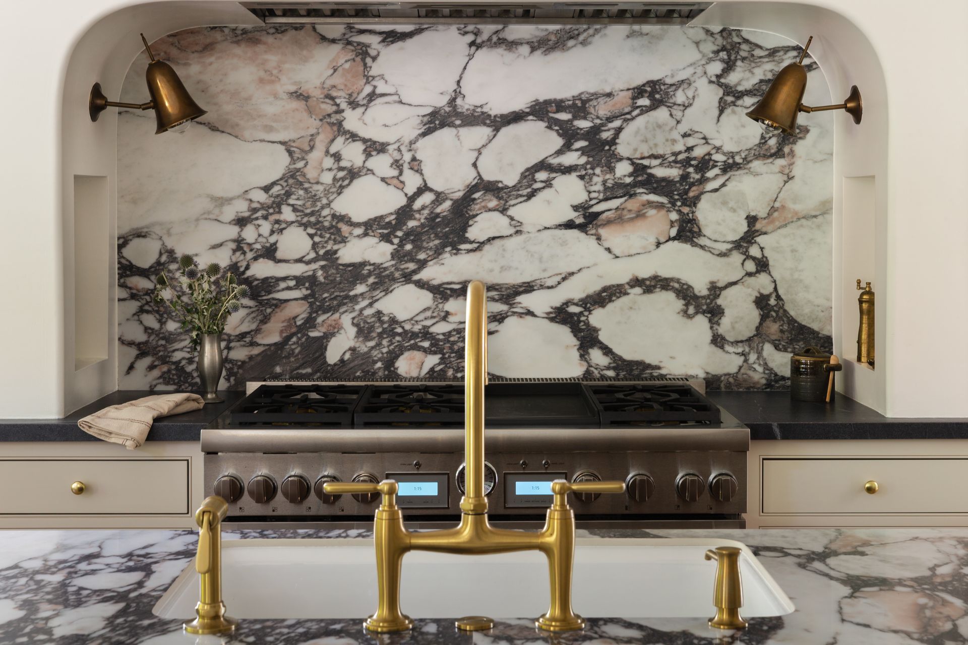 A luxury kitchen with a marble backsplash and countertop, featuring a brass faucet, white farmhouse sink, and gas range.