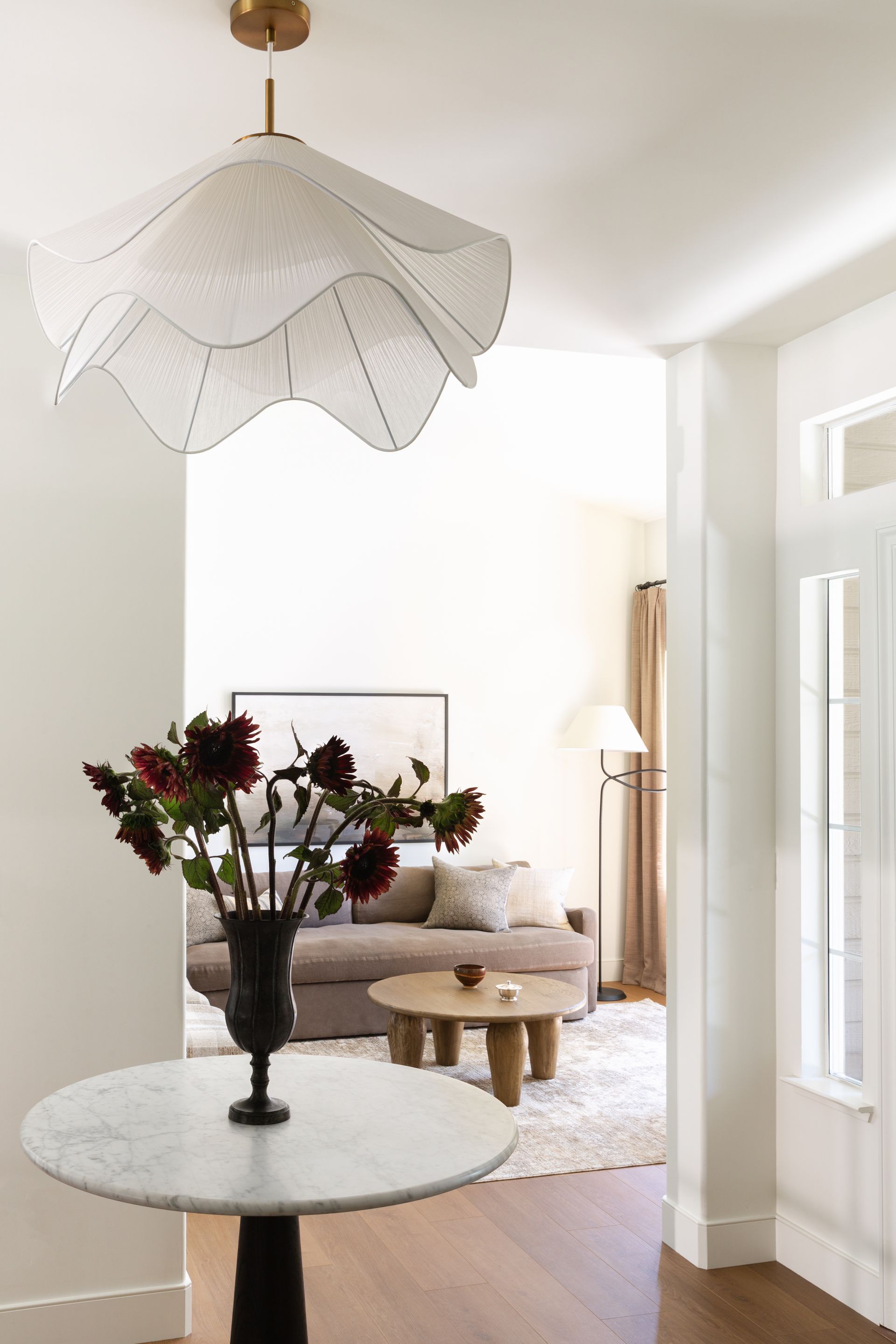A white ruffled pendant light hangs over a marble foyer table with a vase of dark flowers, looking into a living room.