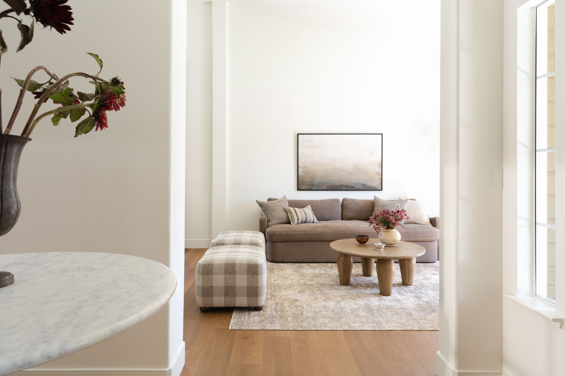 A neutral, modern living room featuring a taupe sofa, plaid ottoman, wooden coffee table, and a large abstract landscape art.
