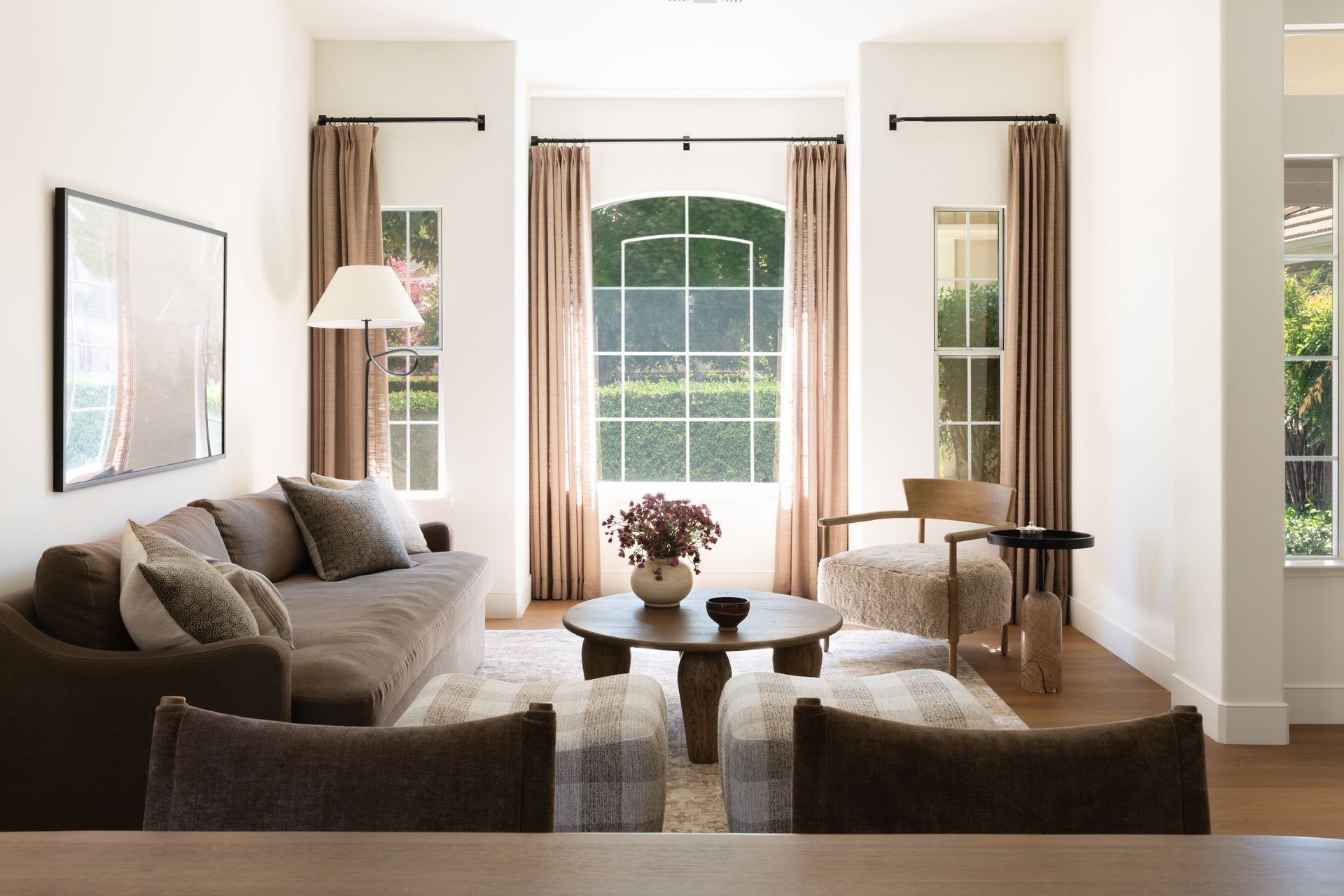 A living room with a brown sofa, a round wooden coffee table, two textured stools, an armchair, and floor-length curtains.