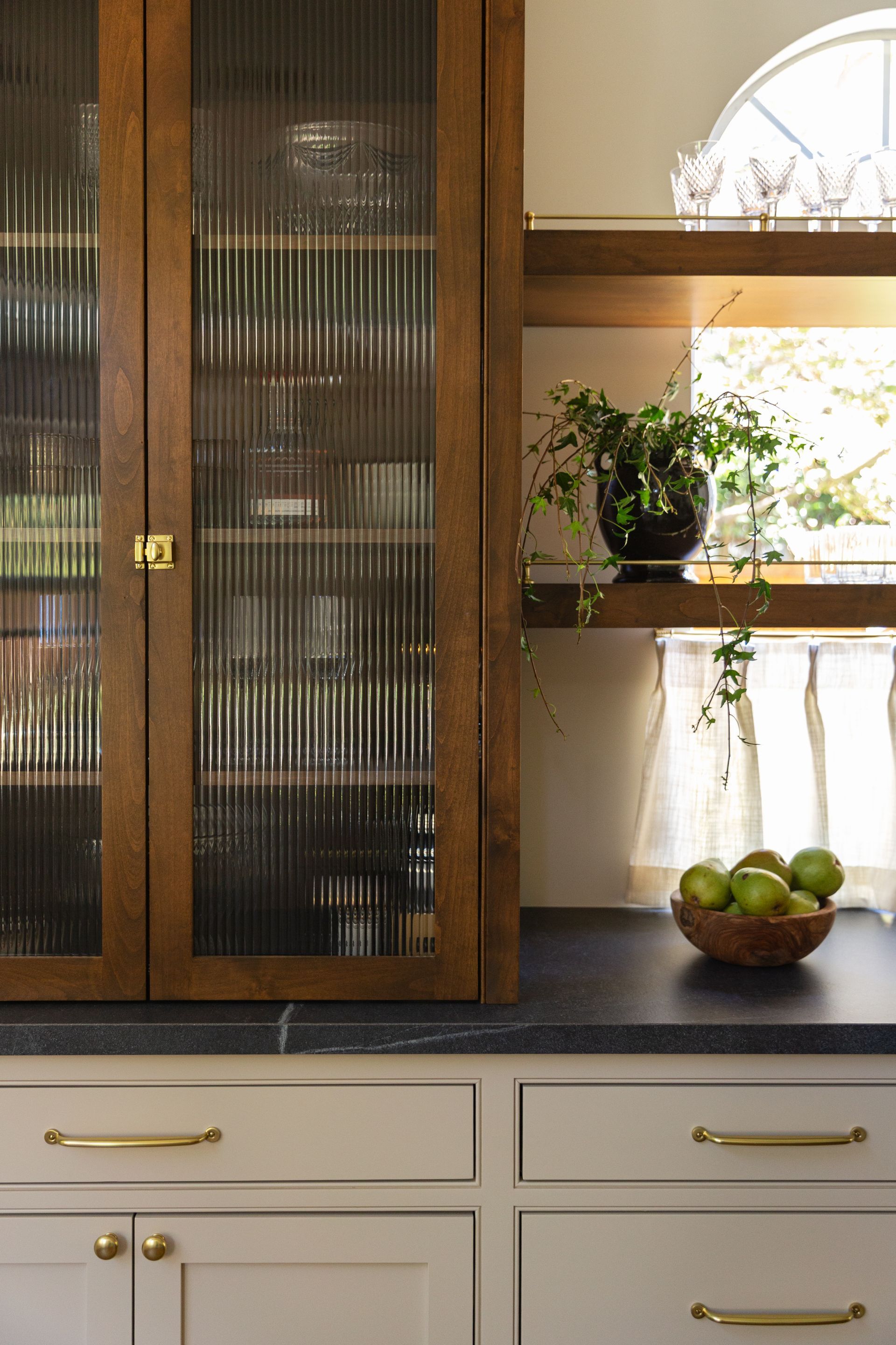A wooden cabinet with reeded glass doors sits next to shelves and light gray cabinets with gold hardware in a kitchen.
