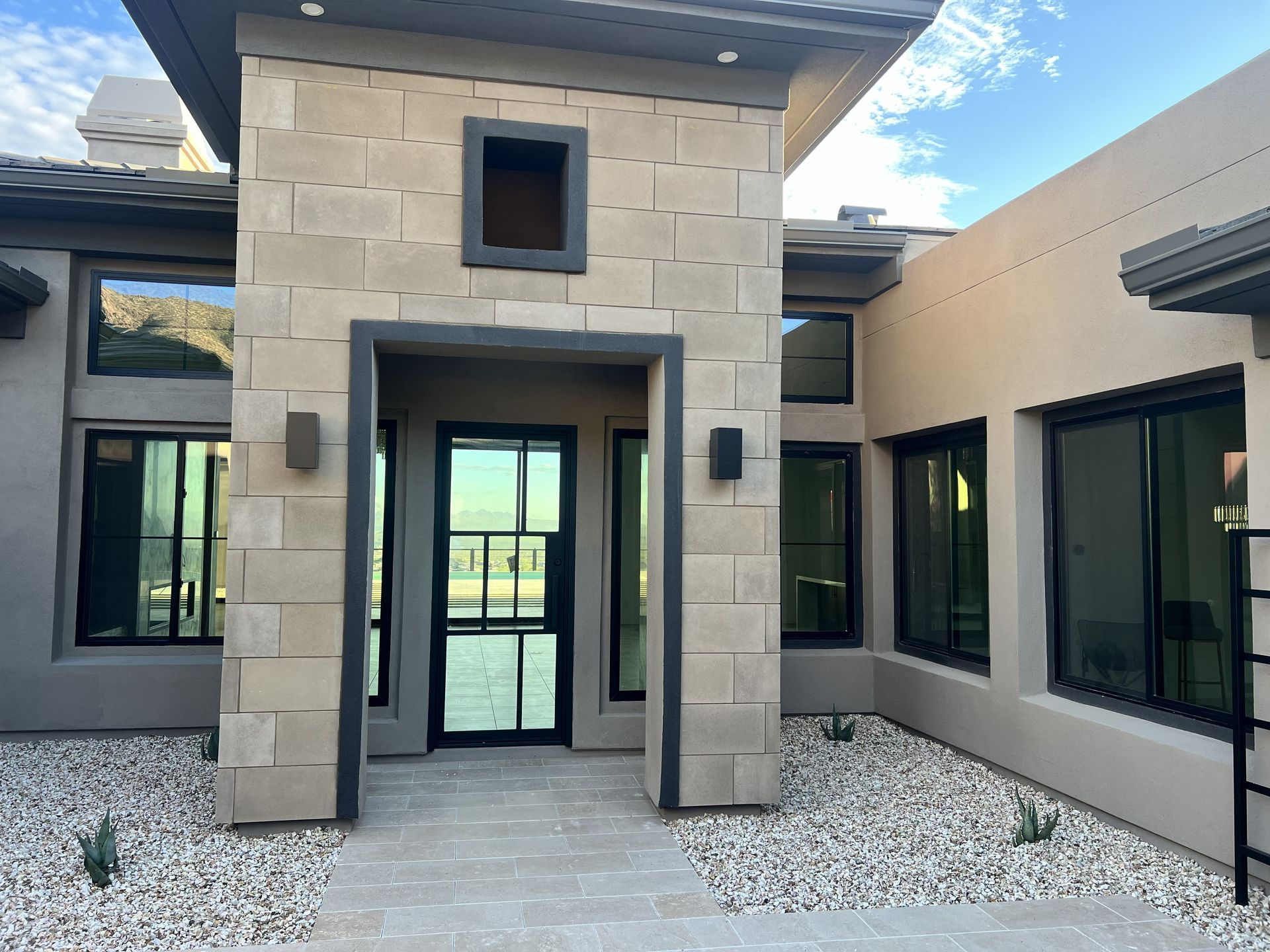 Modern home entryway with stone columns, black-framed windows, and glass double doors over white gravel.