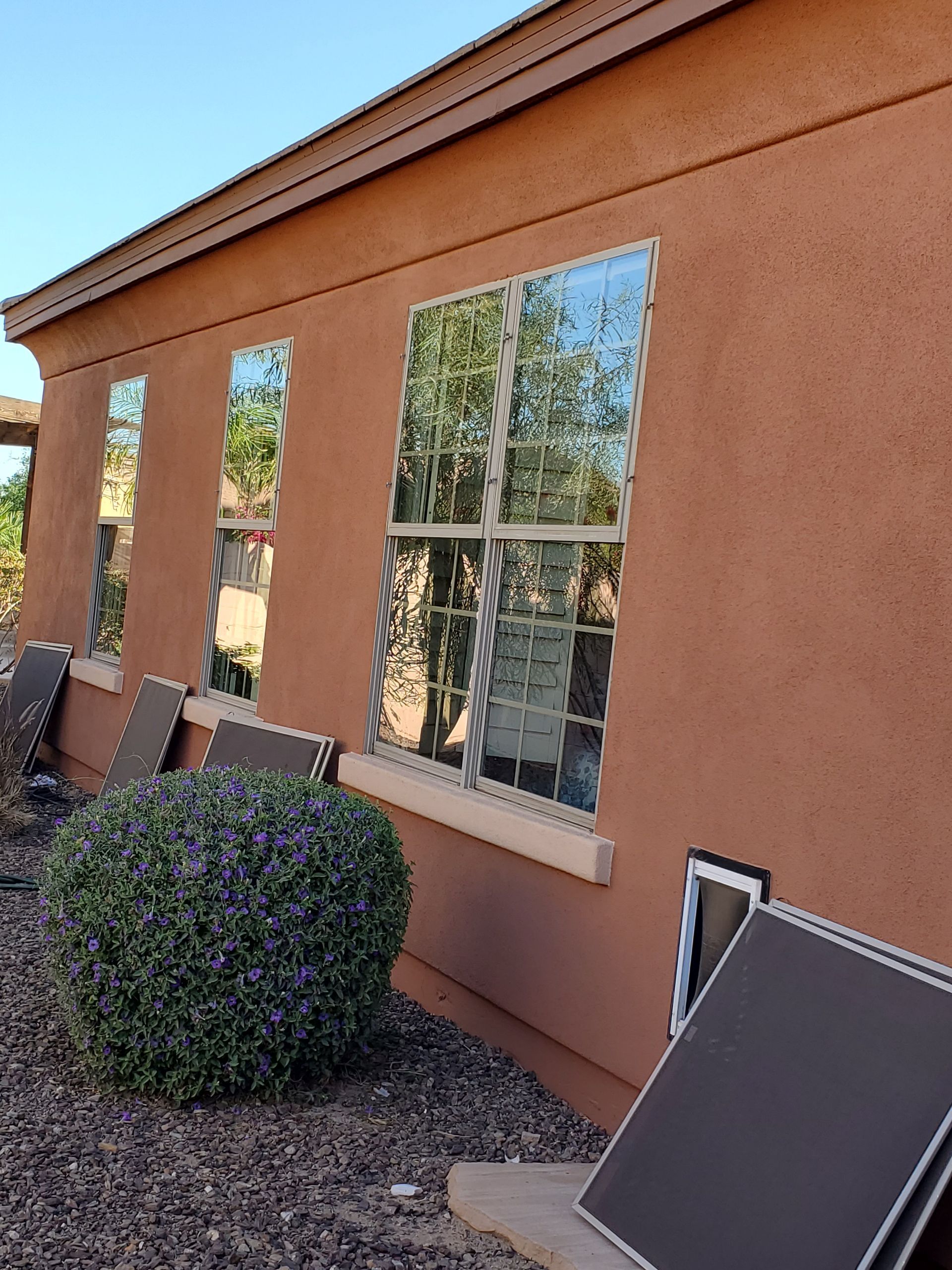 Exterior stucco wall with window reflections, shrubs, and a fallen solar panel in a desert yard