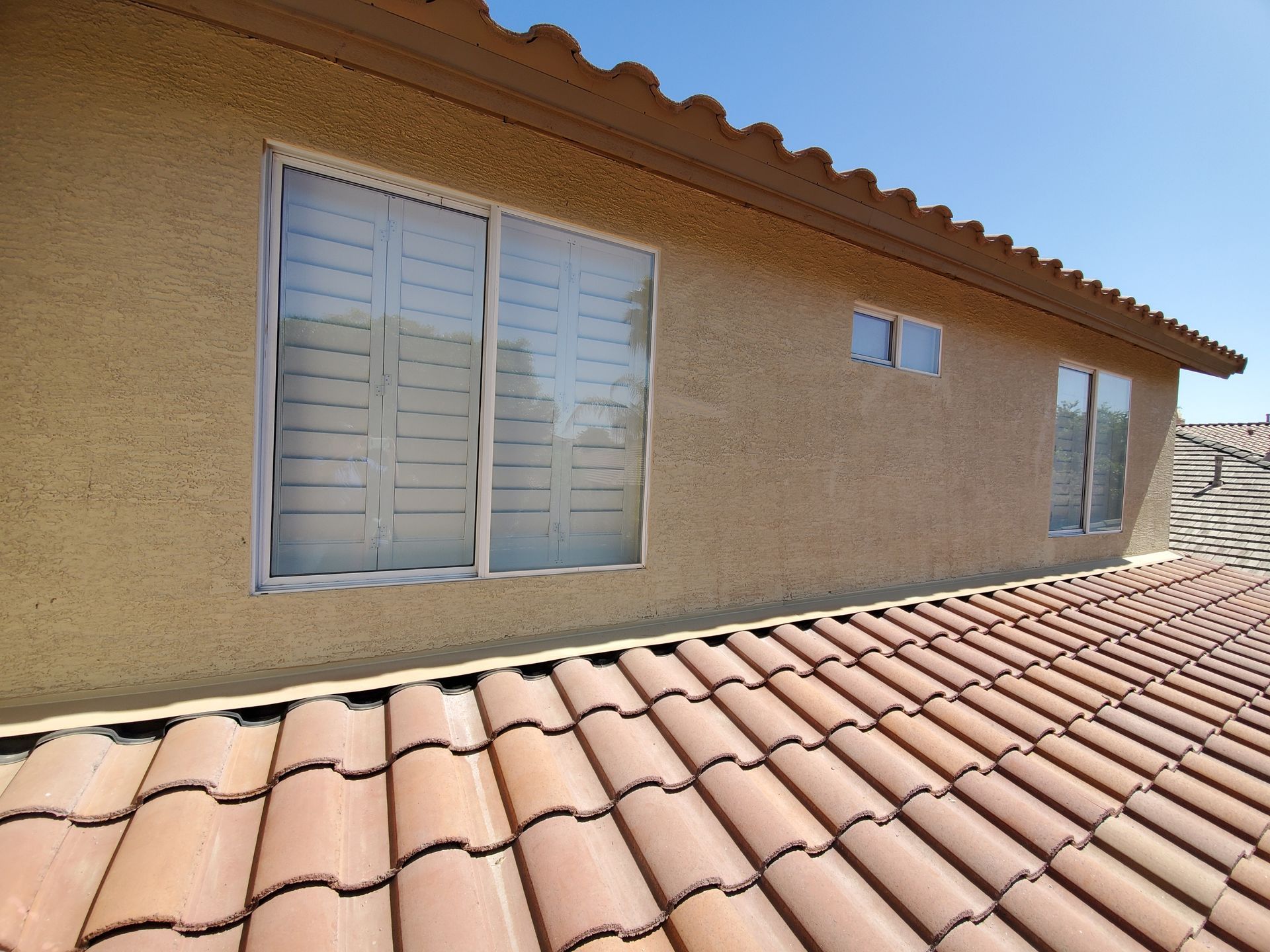 Terracotta roof tiles below a stucco house wall with white windows under a clear blue sky