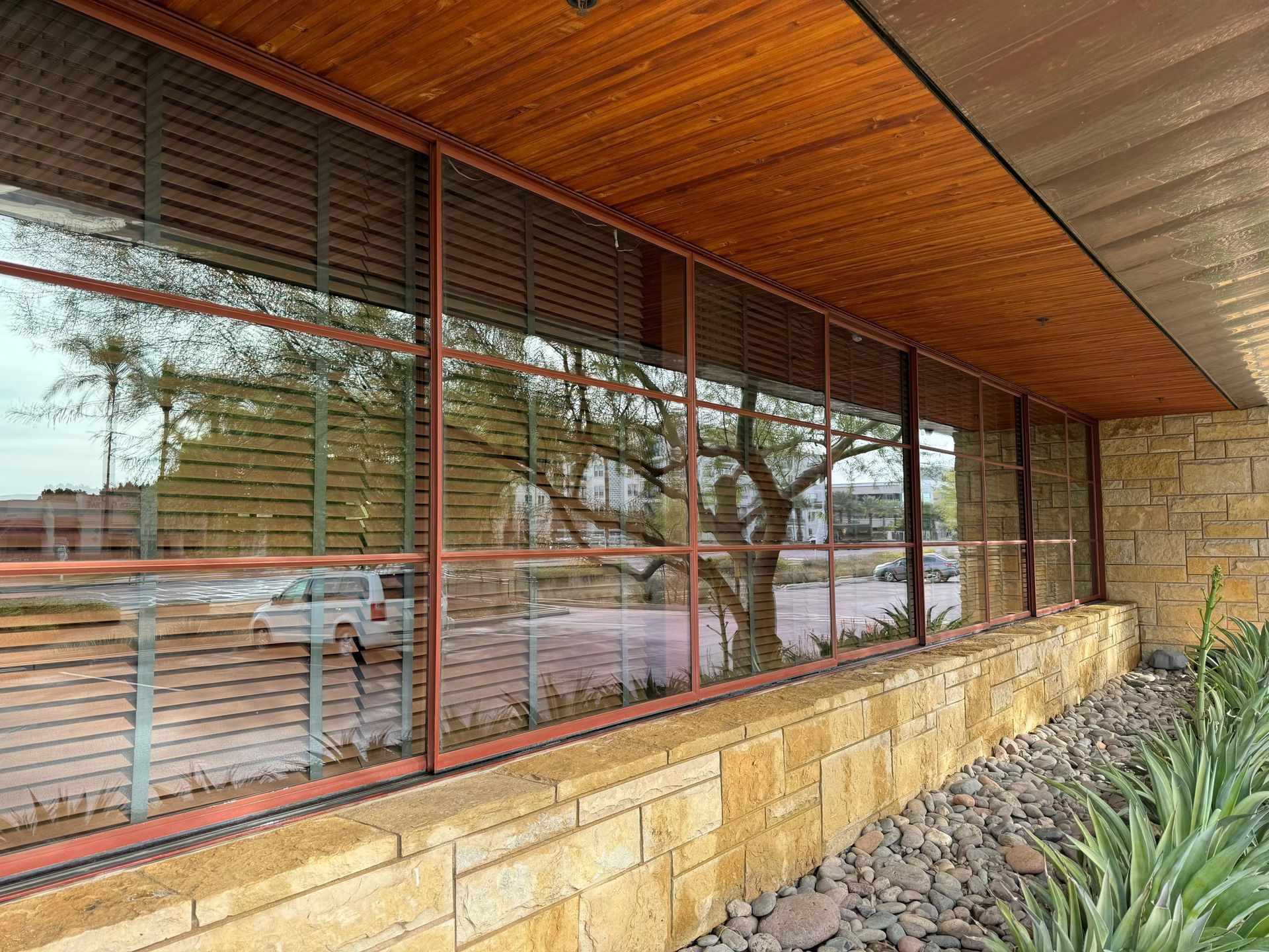Modern building exterior with large windows, wooden overhang, and stone wall along a gravel bed