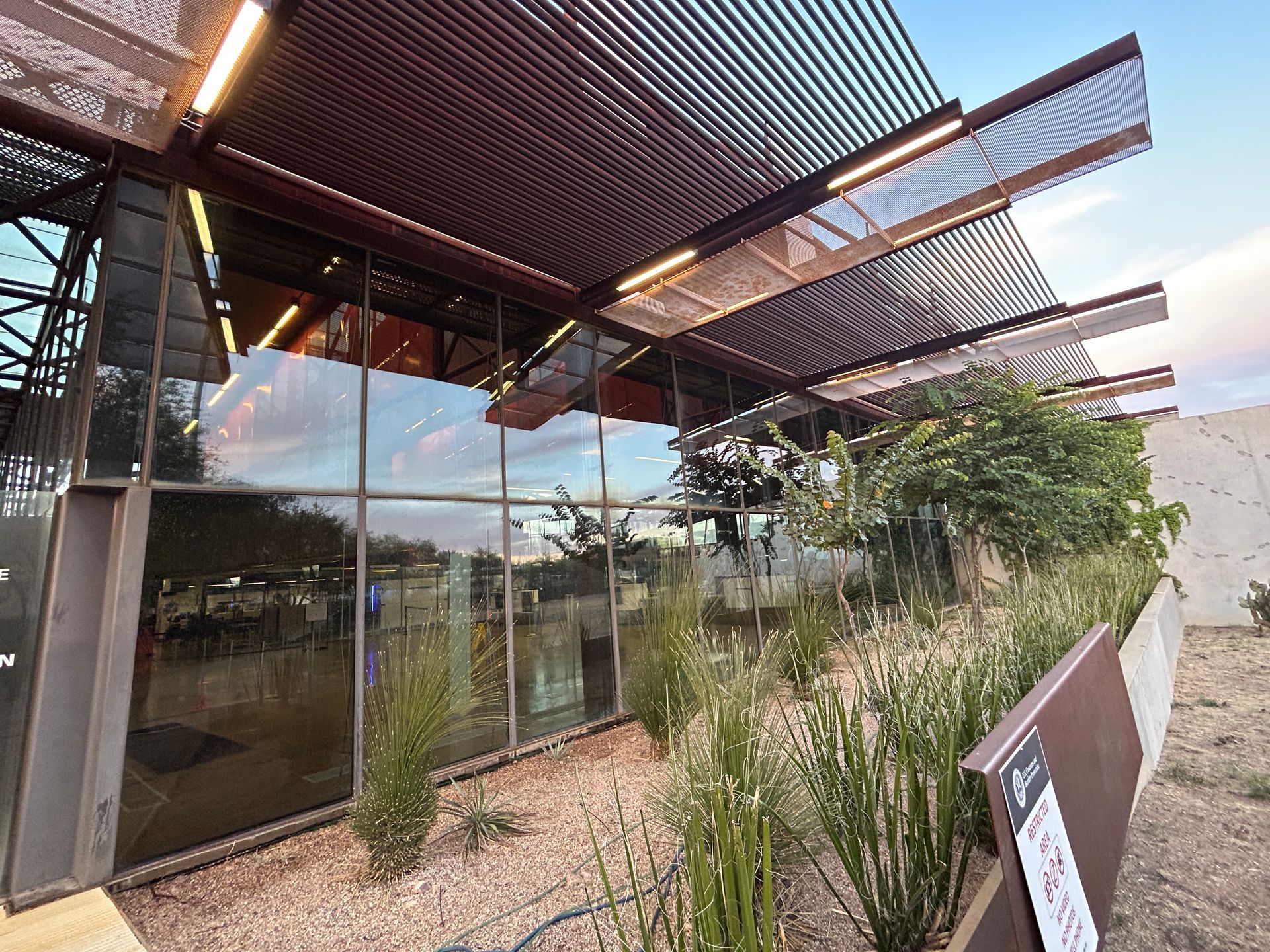 Modern building with glass walls and slatted roof, surrounded by a landscaped garden and gravel path