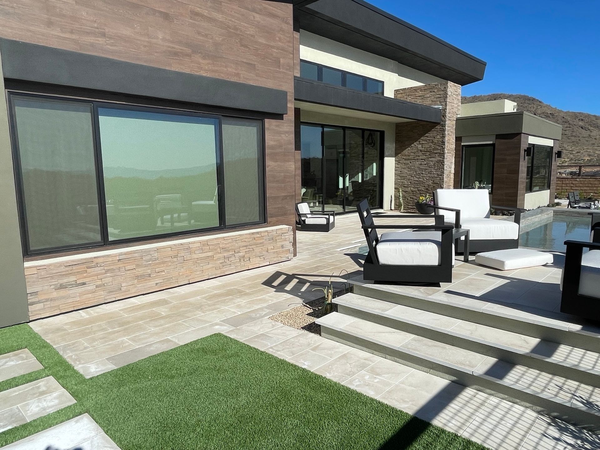 Modern patio with white outdoor seating, large windows, and a green lawn beside a contemporary house